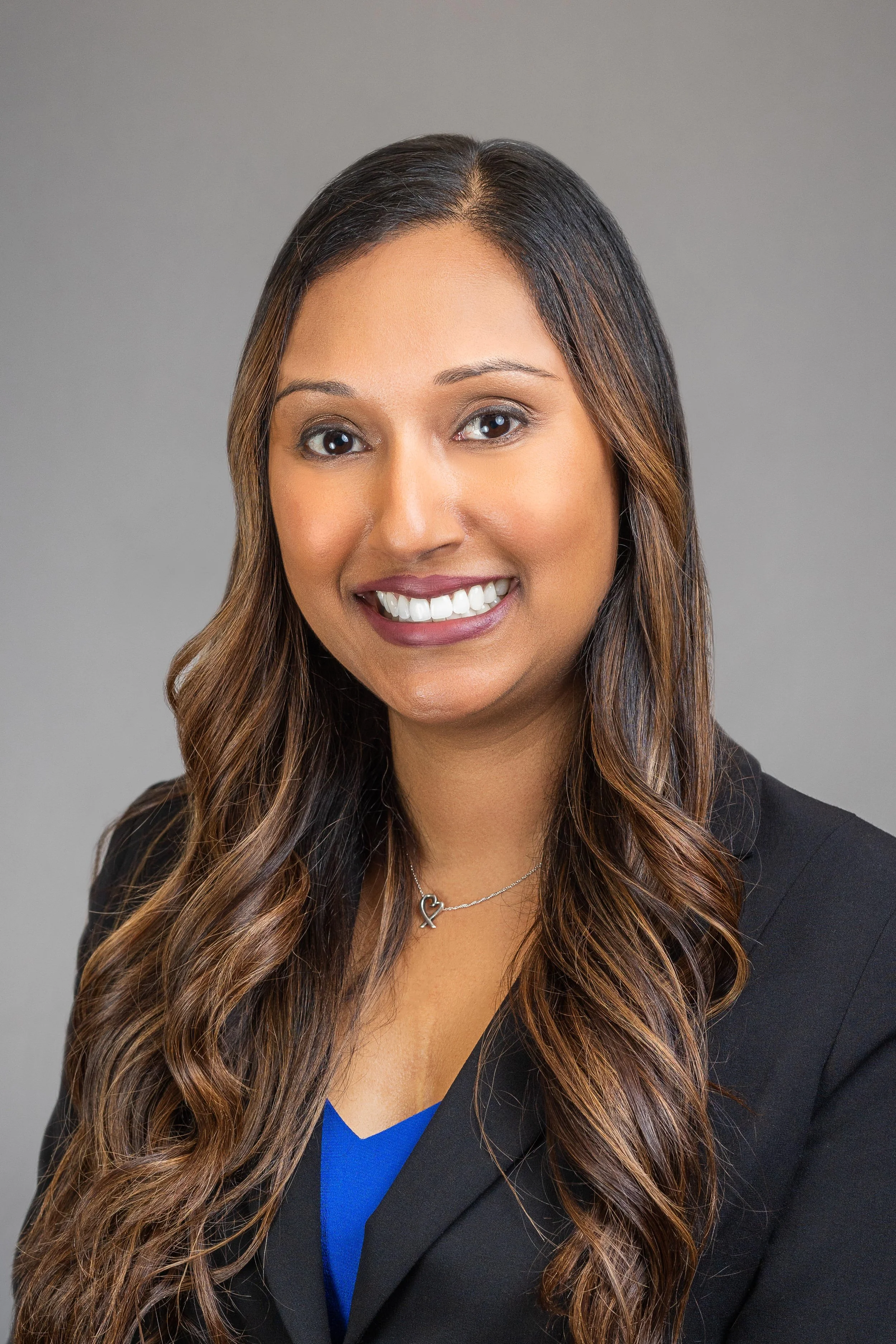 Professional headshot of a smiling woman with long brown hair in loose waves, wearing a black blazer, a blue top, and a silver necklace with a heart-shaped pendant, against a plain gray background.
