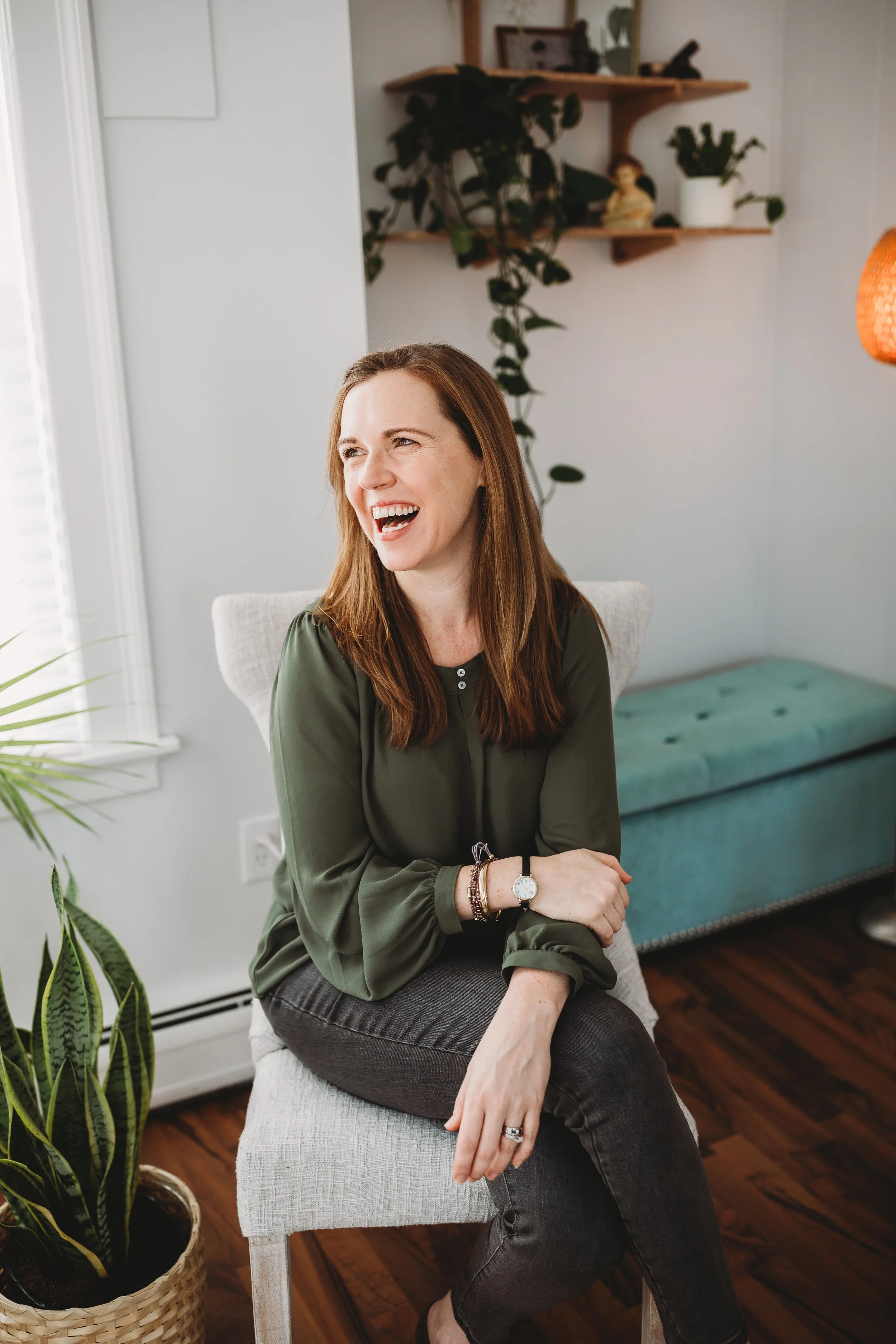A woman with long reddish-brown hair smiling and sitting on a light-colored chair in a living room, wearing a dark green blouse and dark jeans, with a potted plant nearby and decorative shelves in the background.