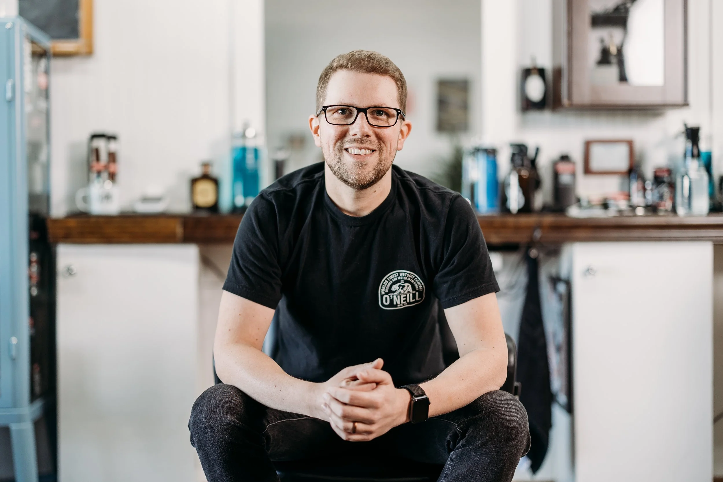 A young man with glasses and a beard, smiling, sitting indoors with a kitchen in the background.