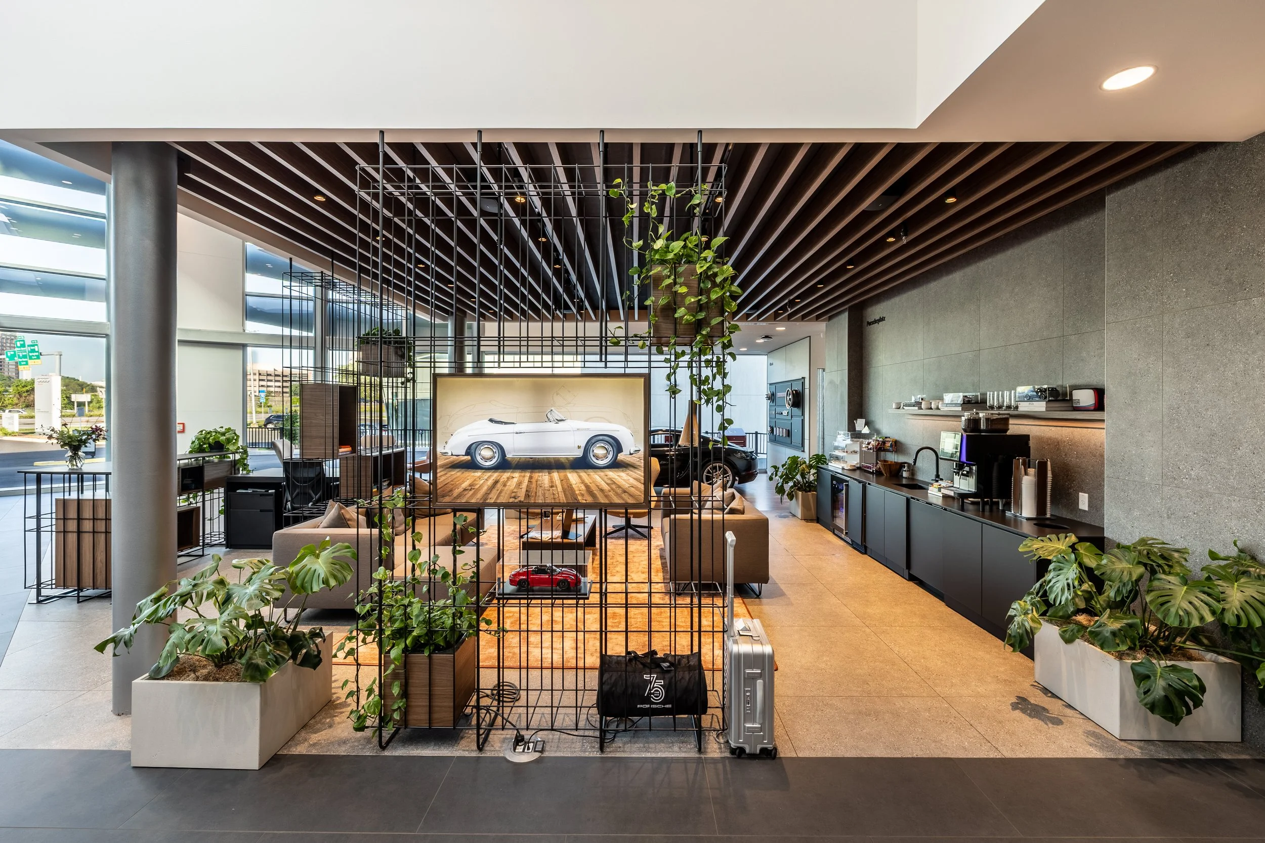 Modern coffee lounge with black cabinets, coffee machine, plants, decorative metal grid, and a large television displaying a white vintage car, accented with wooden ceiling panels.