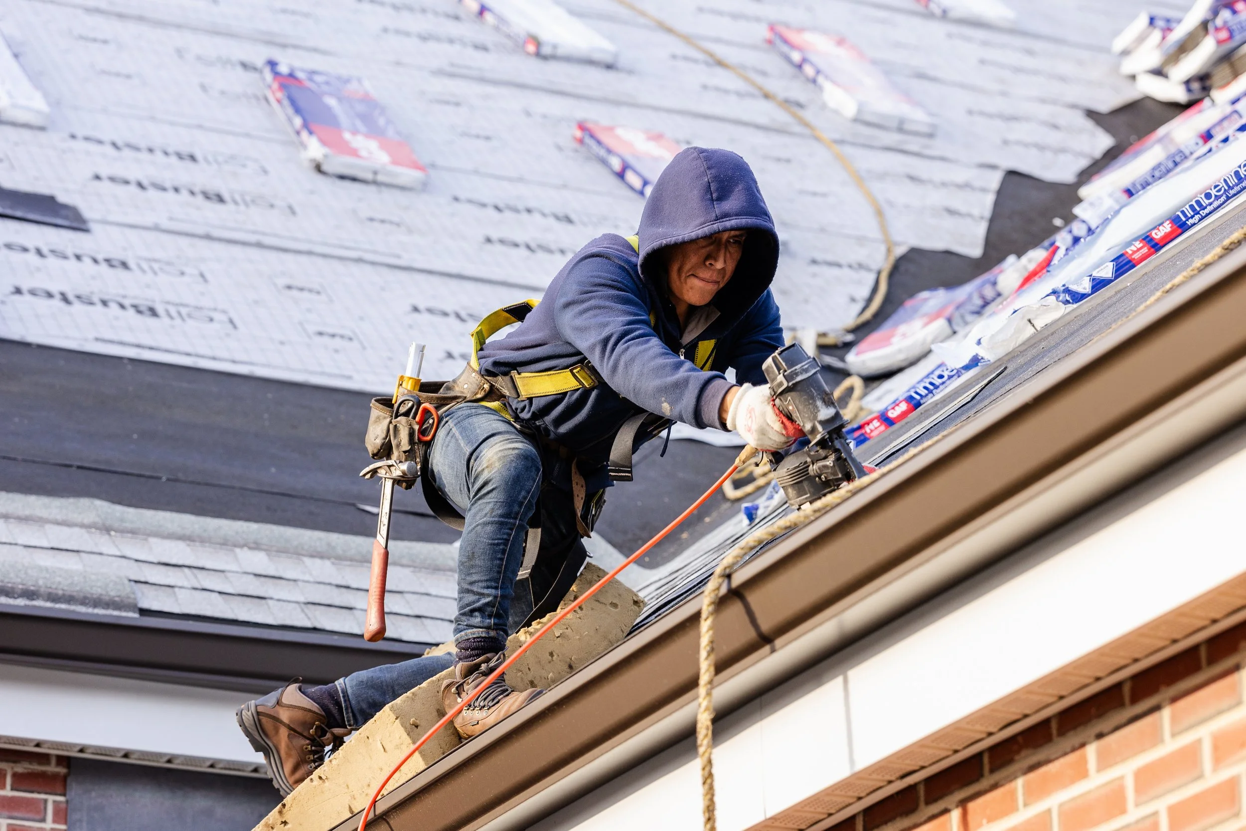 A construction worker wearing a hoodie and safety gloves is installing or repairing roofing shingles on a house. The worker is using a power drill and is perched on a sloped roof with tools hanging from his belt.