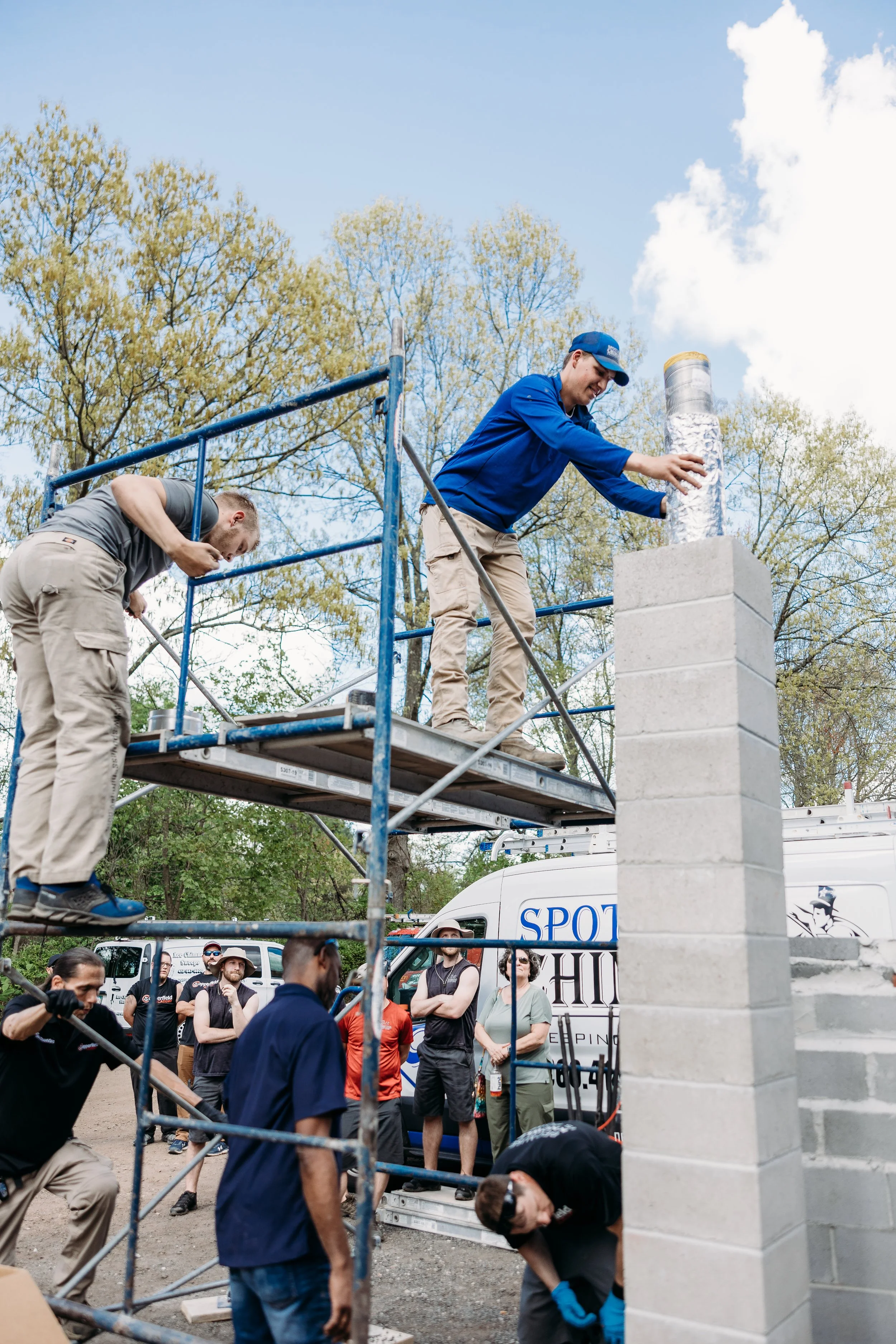 Two men are standing on scaffolding, placing a reflective material on top of a cinder block wall. Several people are watching nearby, and there are trucks and trees in the background with a partly cloudy sky.