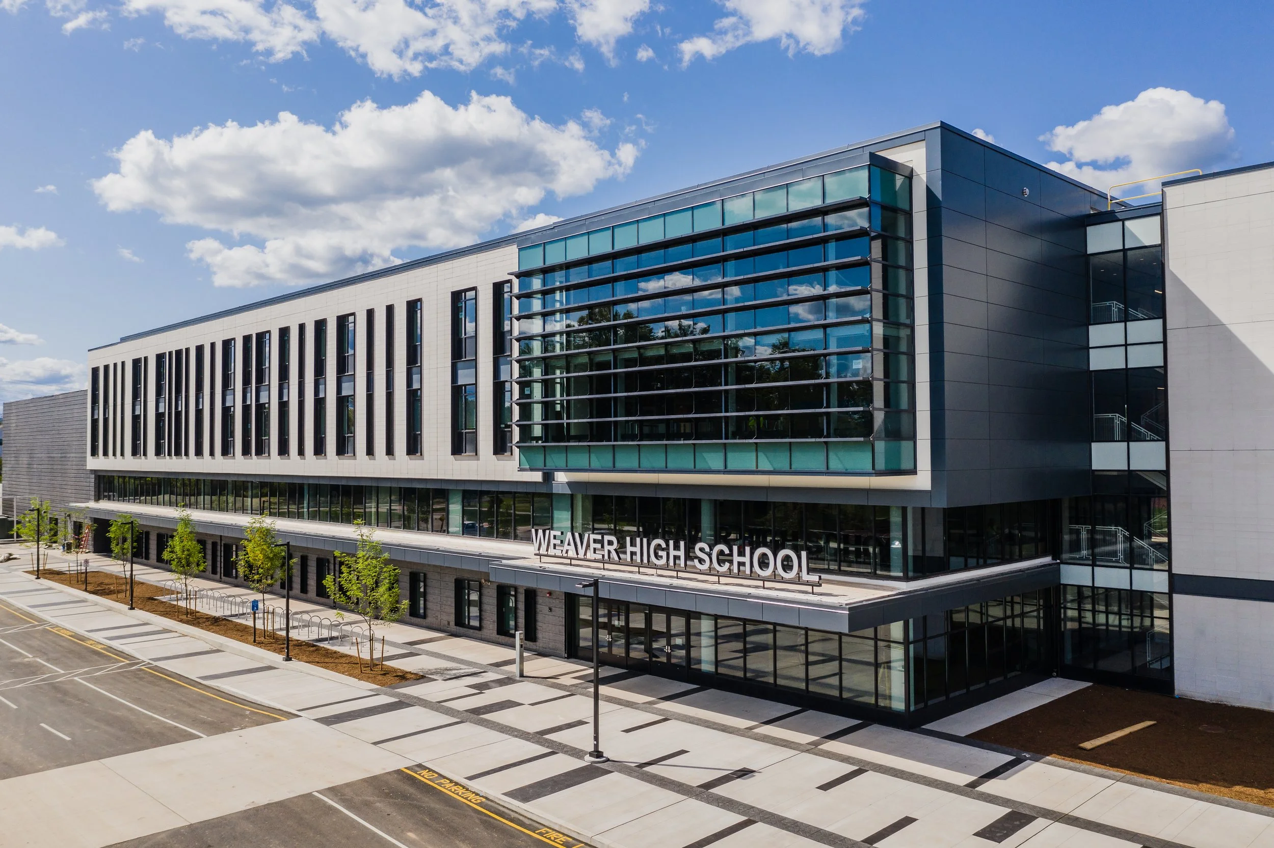 Exterior view of Weaver High School building with glass windows and trees lining the sidewalk on a clear, sunny day.