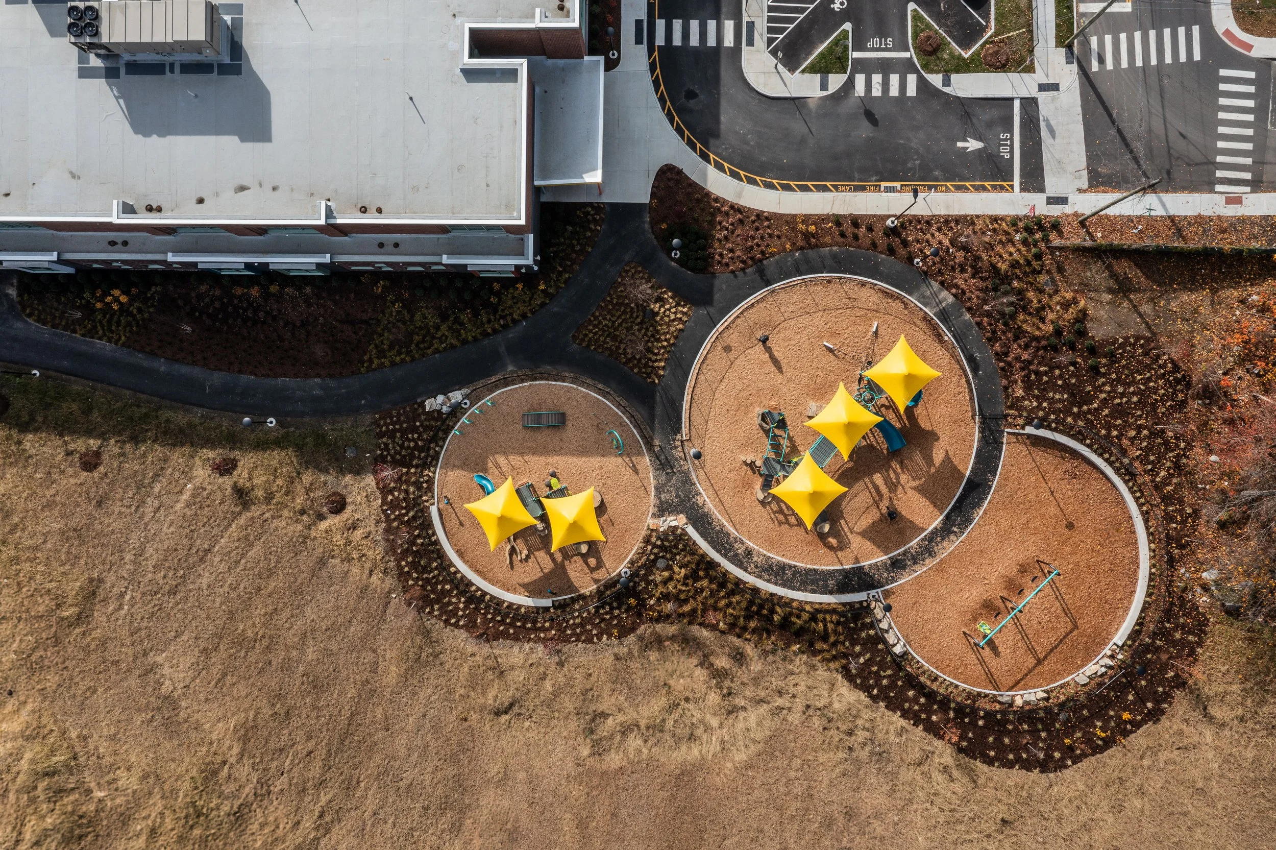 An aerial view of a small playground with yellow shade tents, slides, and swings, situated near a building and parking lot in a park area.
