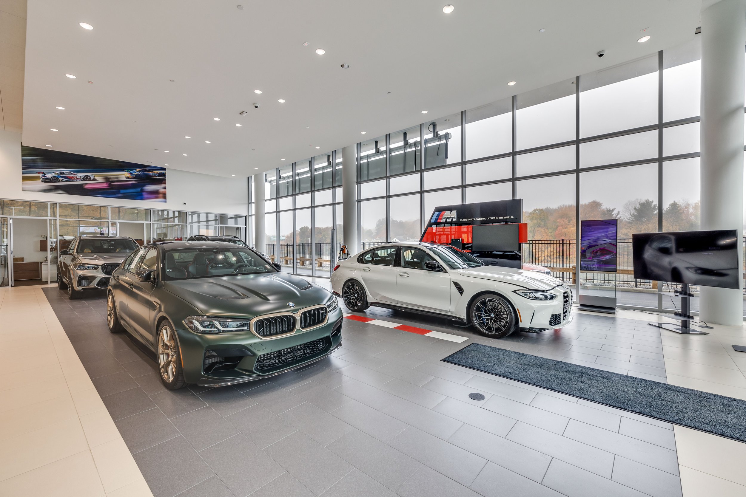 Showroom with several luxury cars on display, including a matte green BMW, a white sports car, and another vehicle in the background, with large windows and bright lighting.