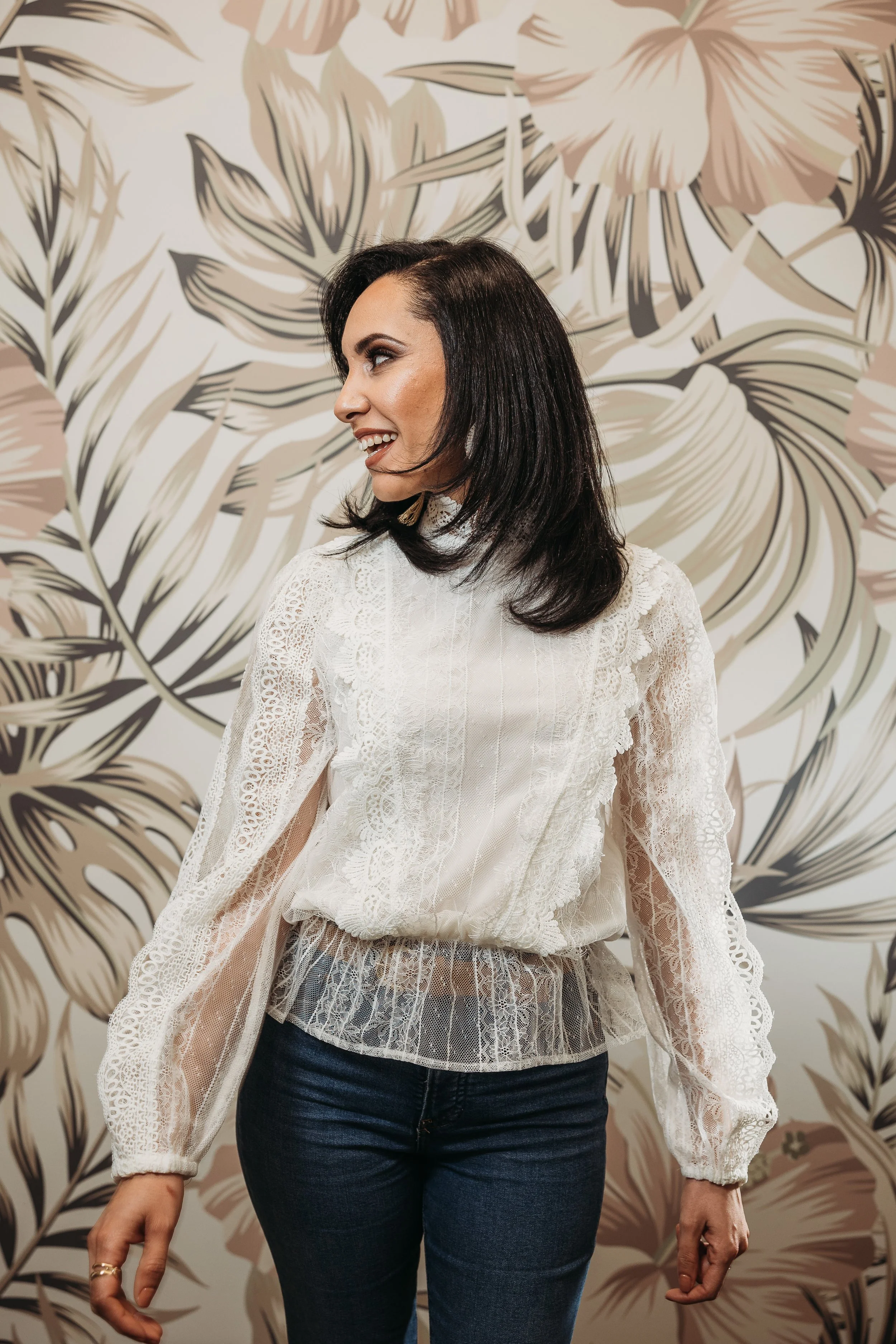 A woman with dark hair wearing a white lace blouse and dark jeans standing in front of a floral wallpaper background, smiling and looking to her left.