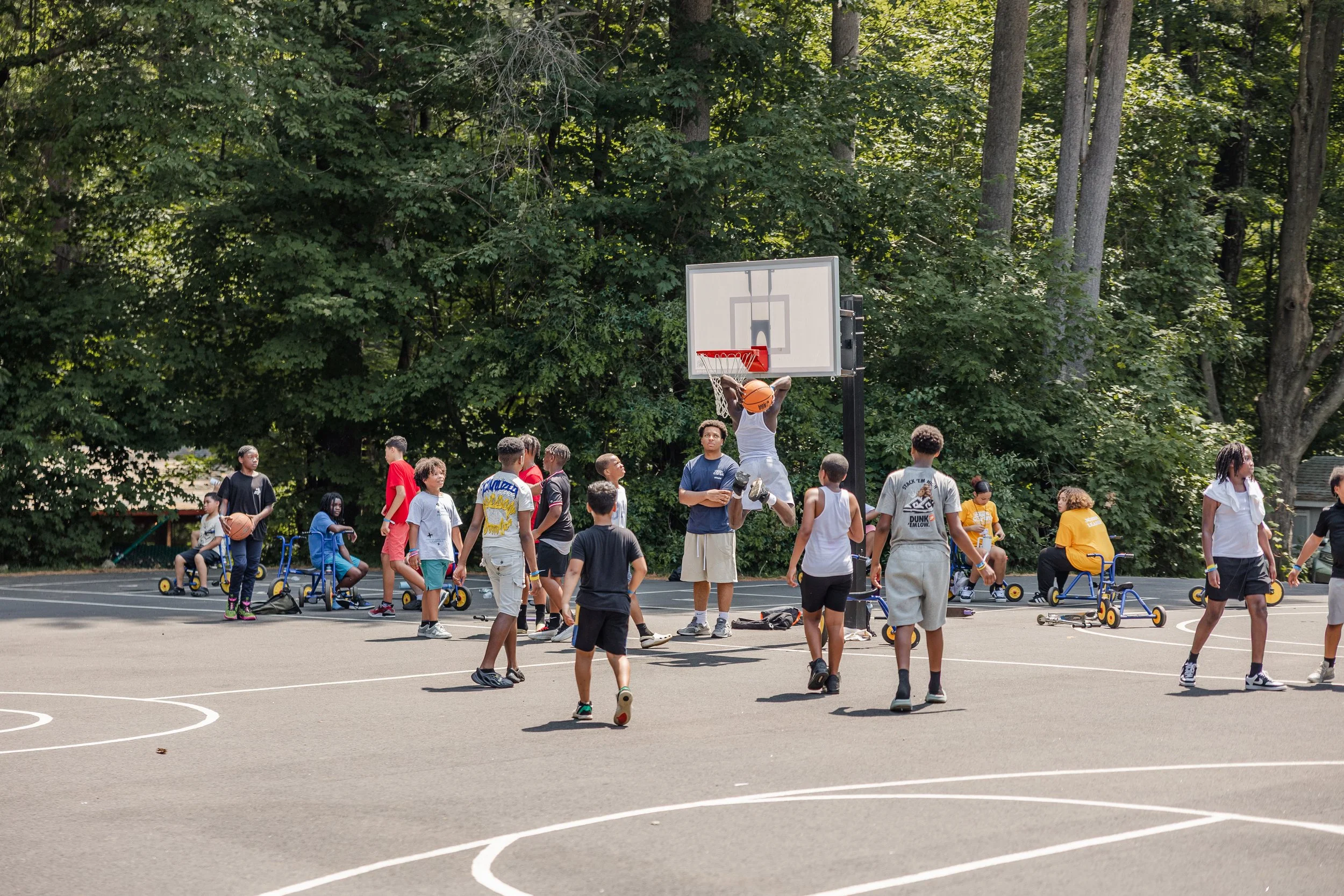 Children playing basketball on an outdoor court surrounded by tall trees.