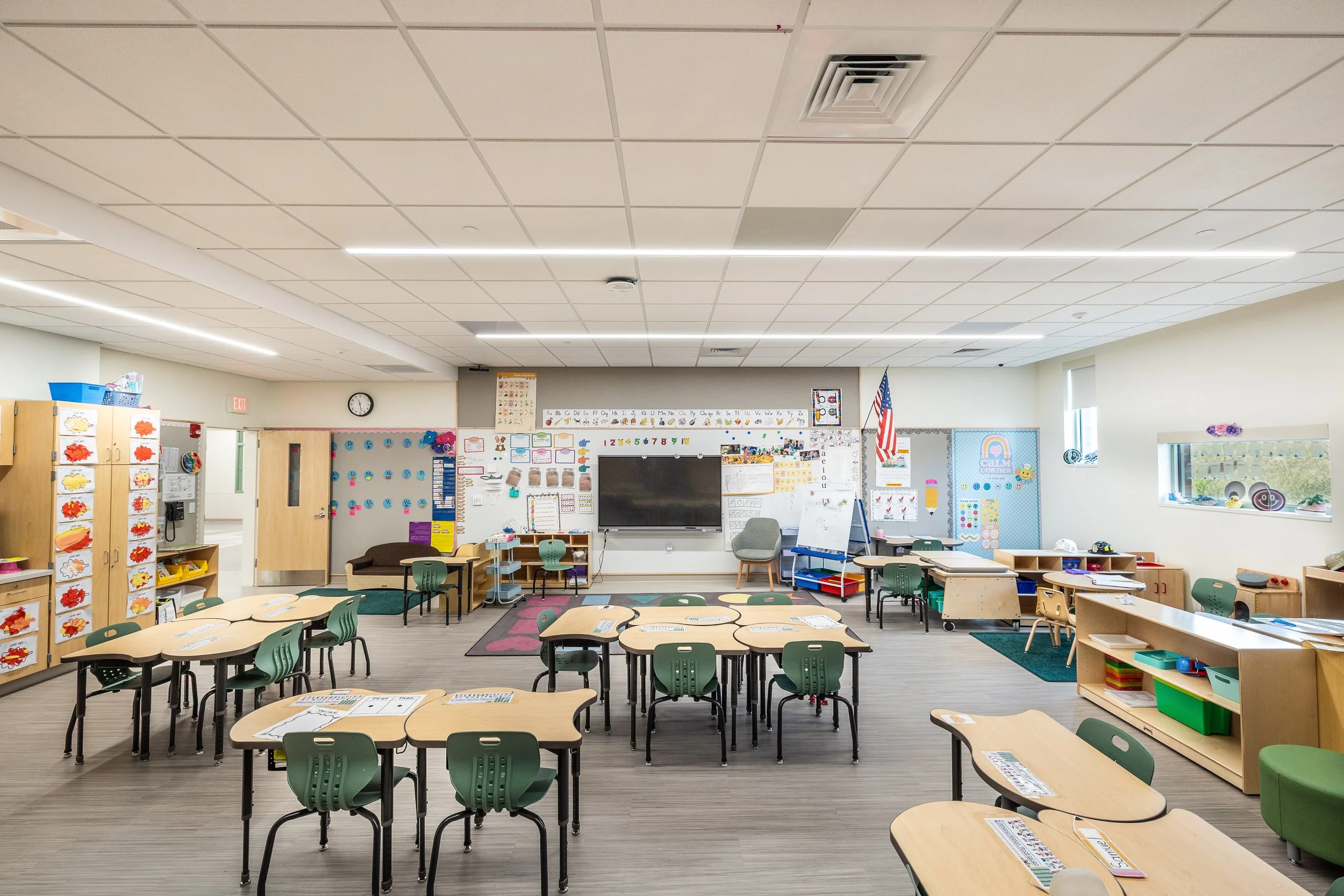 Empty kindergarten classroom with desks, chairs, storage cabinets, educational materials, and colorful decorations on the walls.