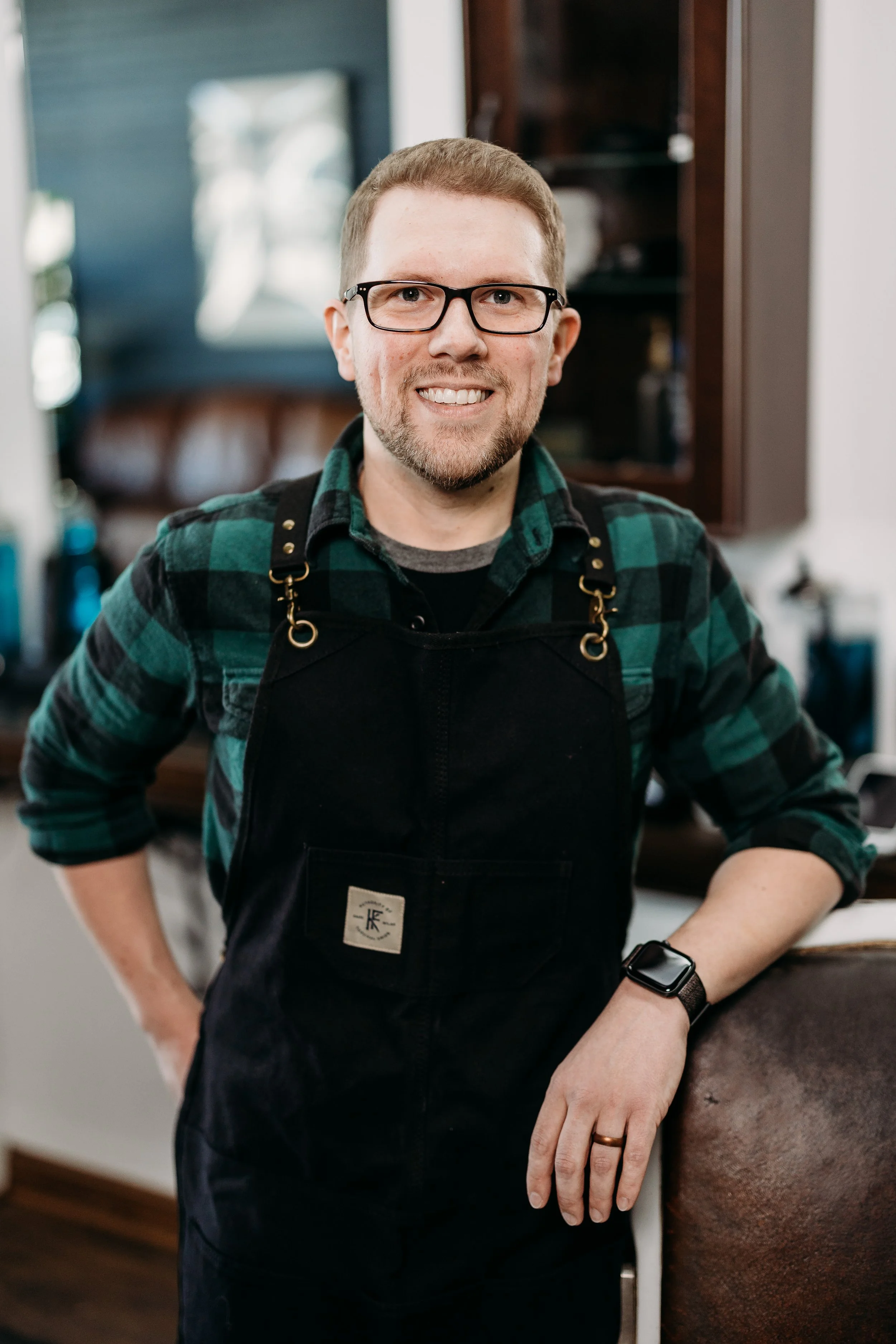 A man with glasses and a beard wearing a green plaid shirt and black apron, standing in a kitchen.