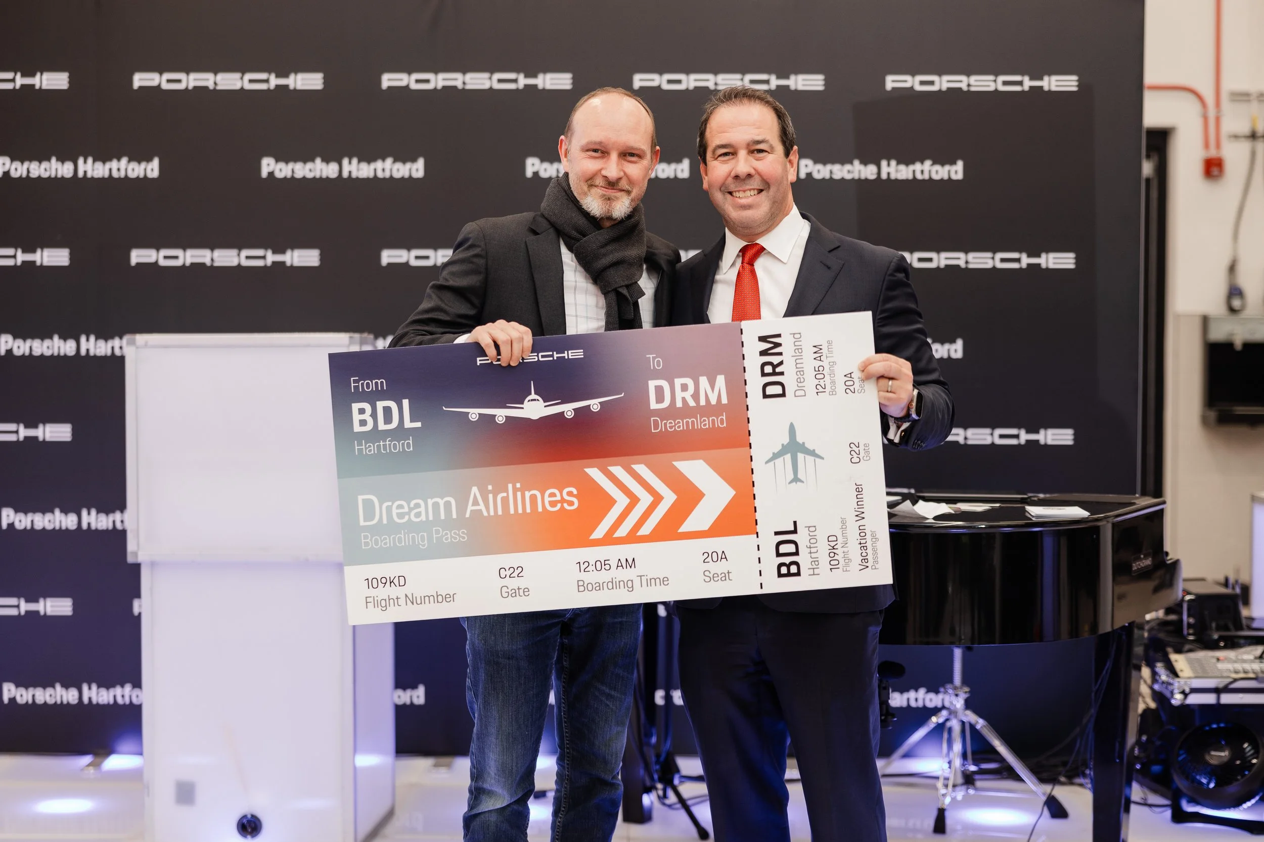 Two men in business suits holding a large mock airline boarding pass at a Porsche Hartford event, with a backdrop marking Porsche branding.