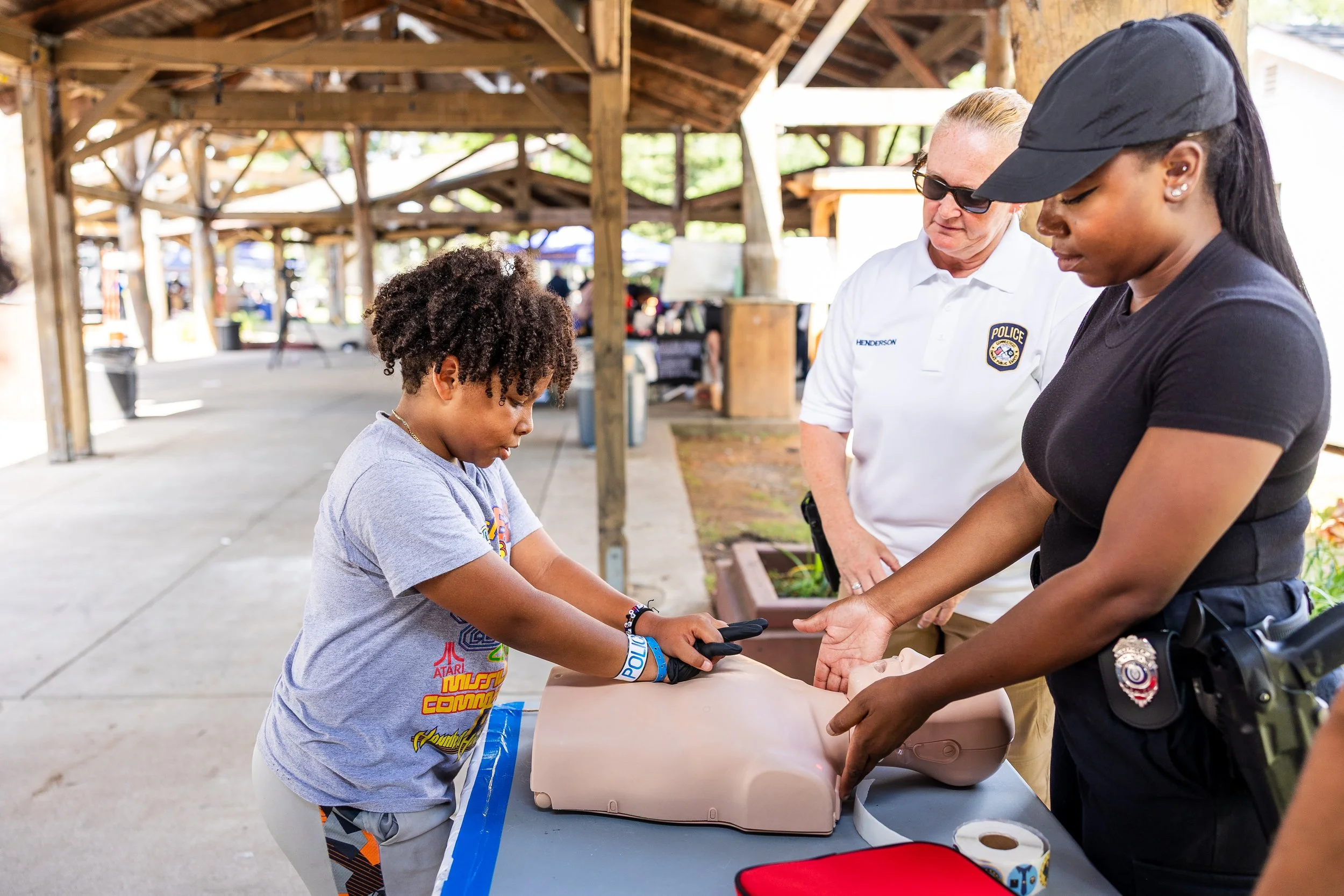A young boy learning CPR on a training mannequin at an outdoor event, overseen by two female police officers.