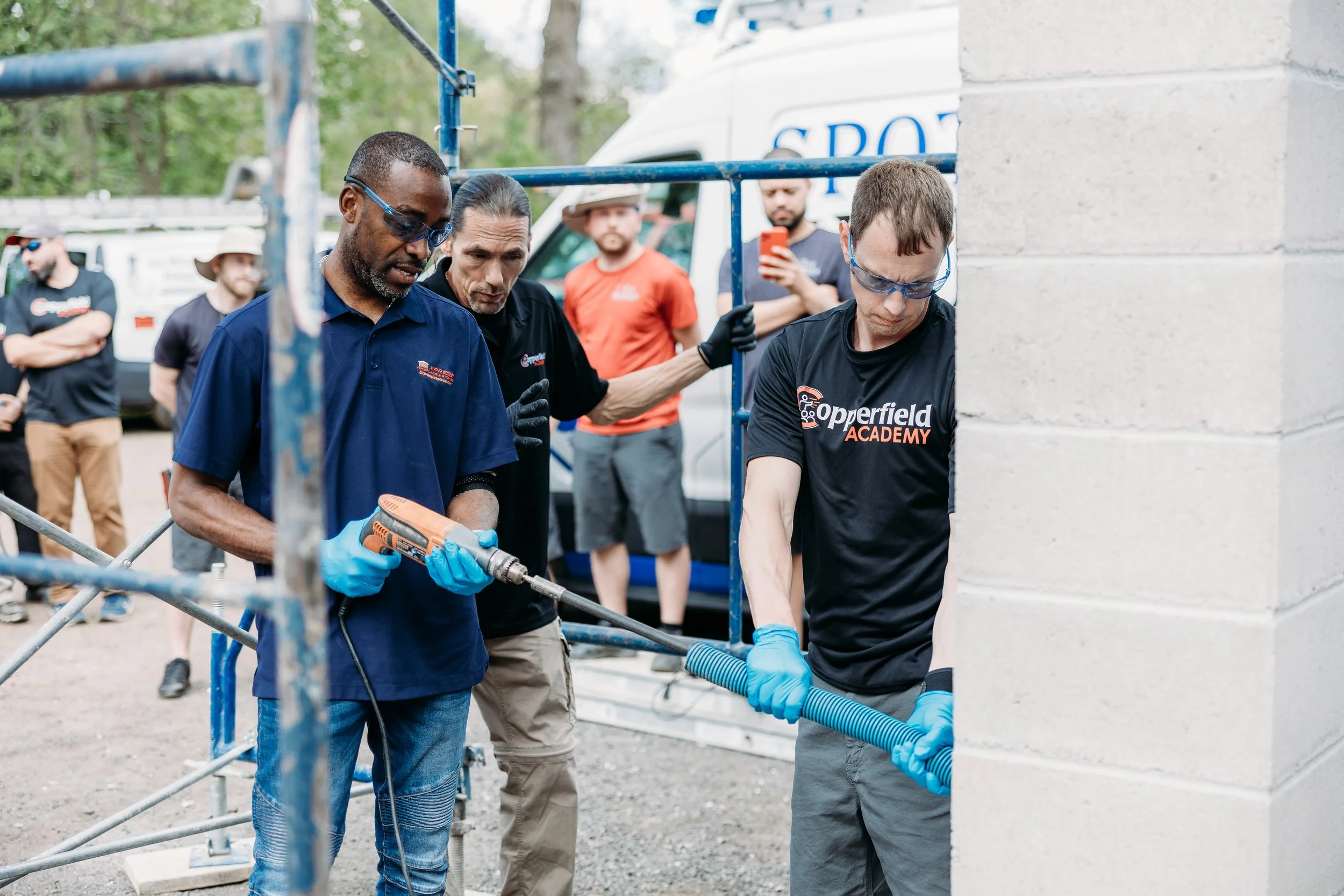 Three men wearing safety glasses and gloves working with a large drill and a flexible pipe or hose, in an outdoor construction or training site with onlookers and a van in the background.