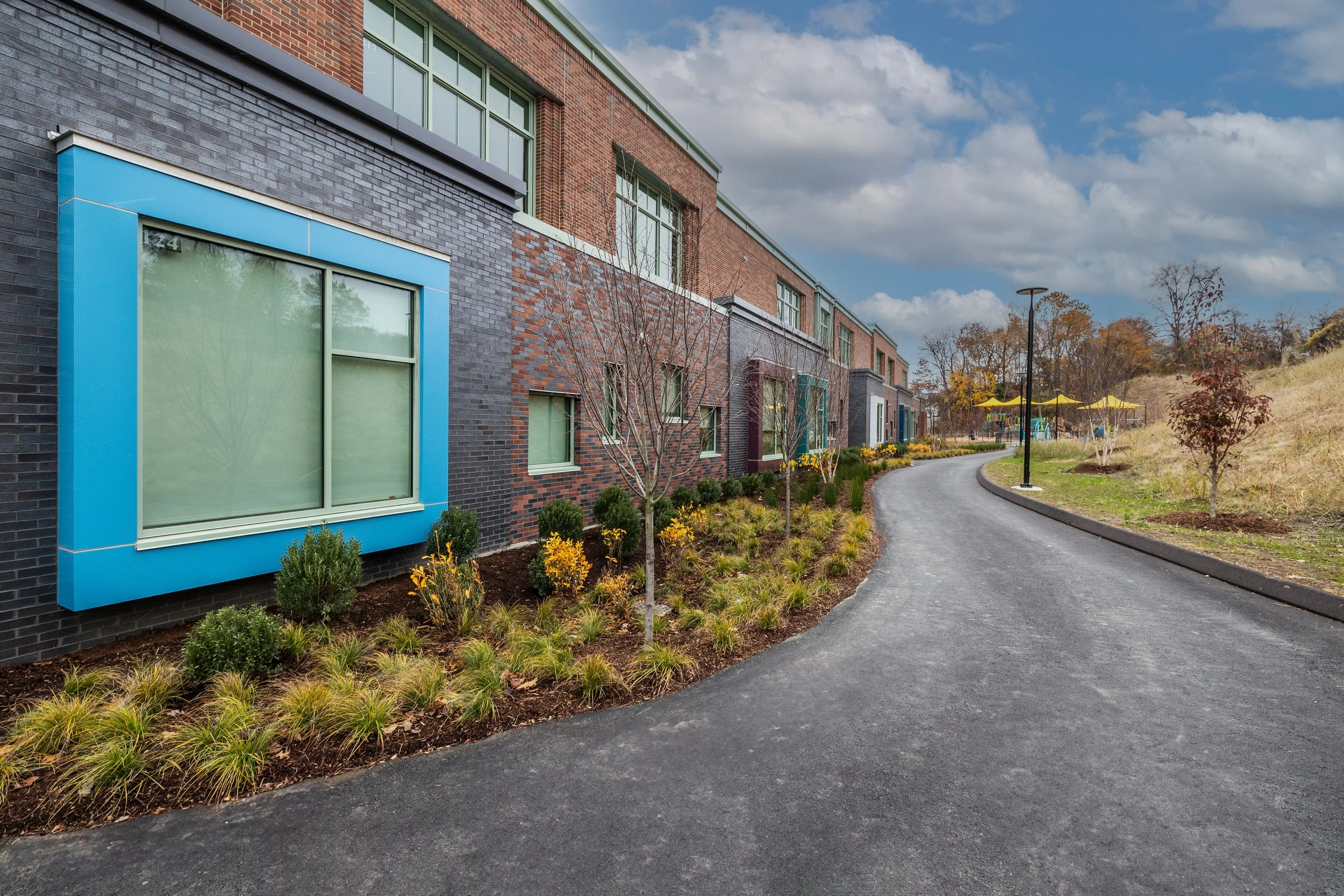 A paved sidewalk running alongside a modern multi-story building with colorful window frames, surrounded by landscaped plants and small trees, with an outdoor seating area with yellow umbrellas in the background under a partly cloudy sky.