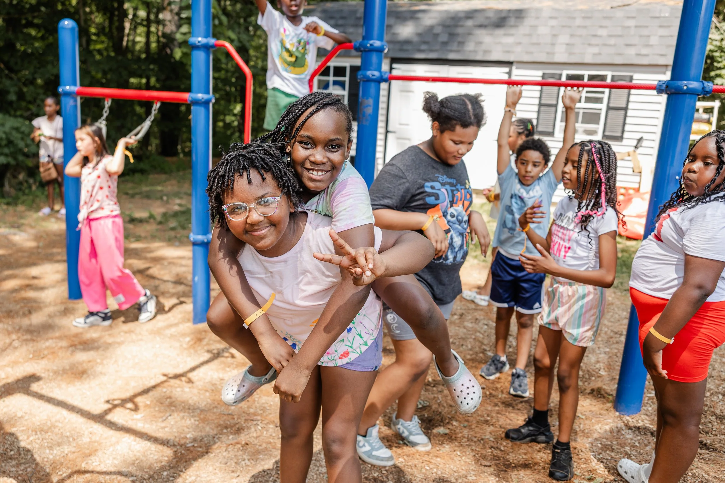 Children playing on a playground, with two girls in the foreground smiling and piggybacking, surrounded by other kids.