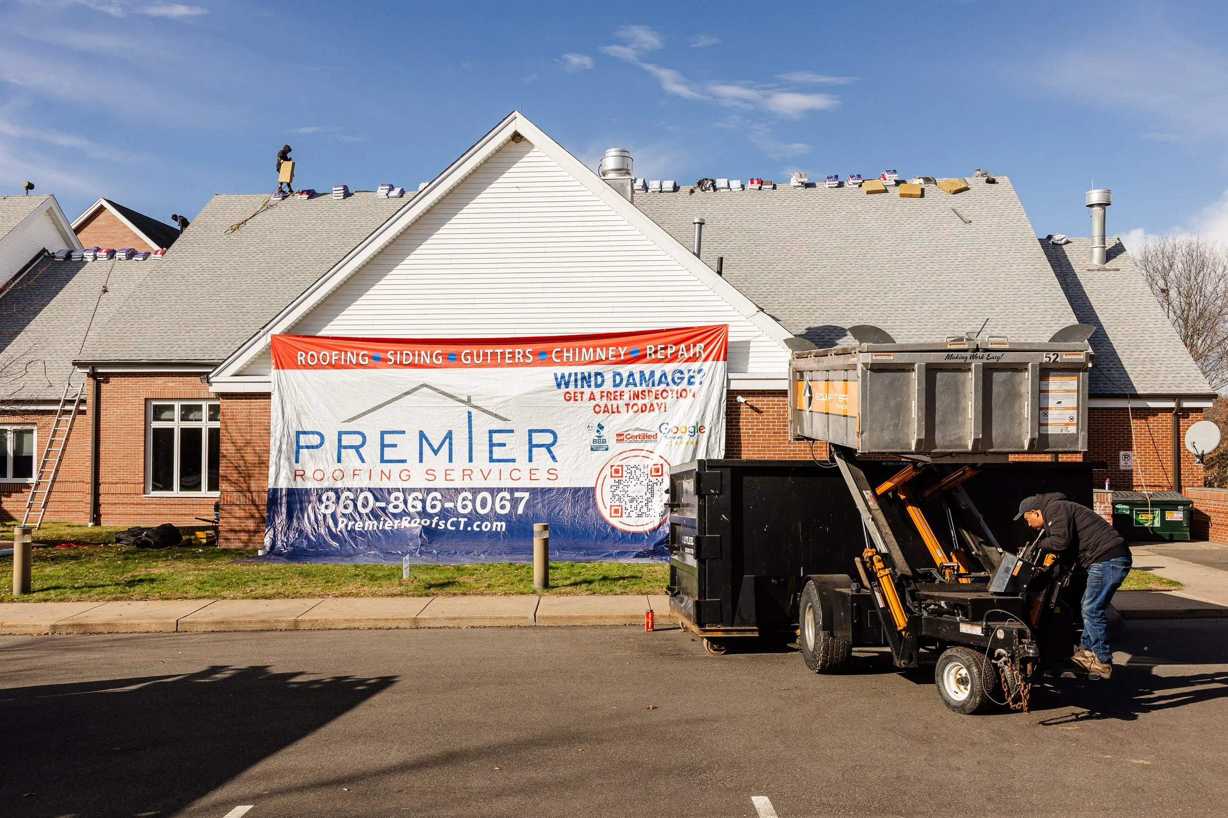 A house with workers installing a roof, with shingles and a banner advertising Premier Roofing Services on the front.