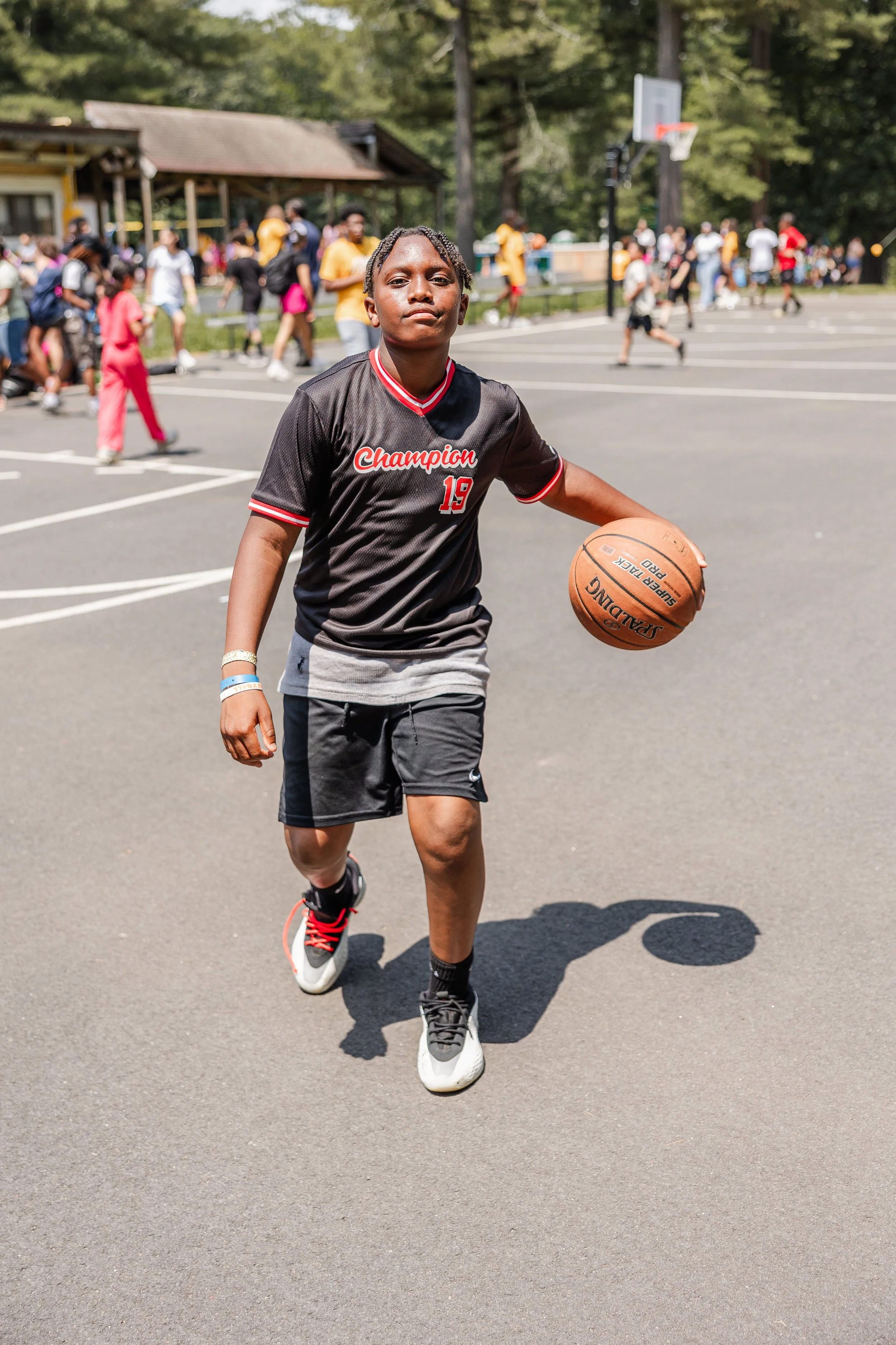 A young boy dribbling a basketball on an outdoor basketball court, with a crowd of people walking in the background.