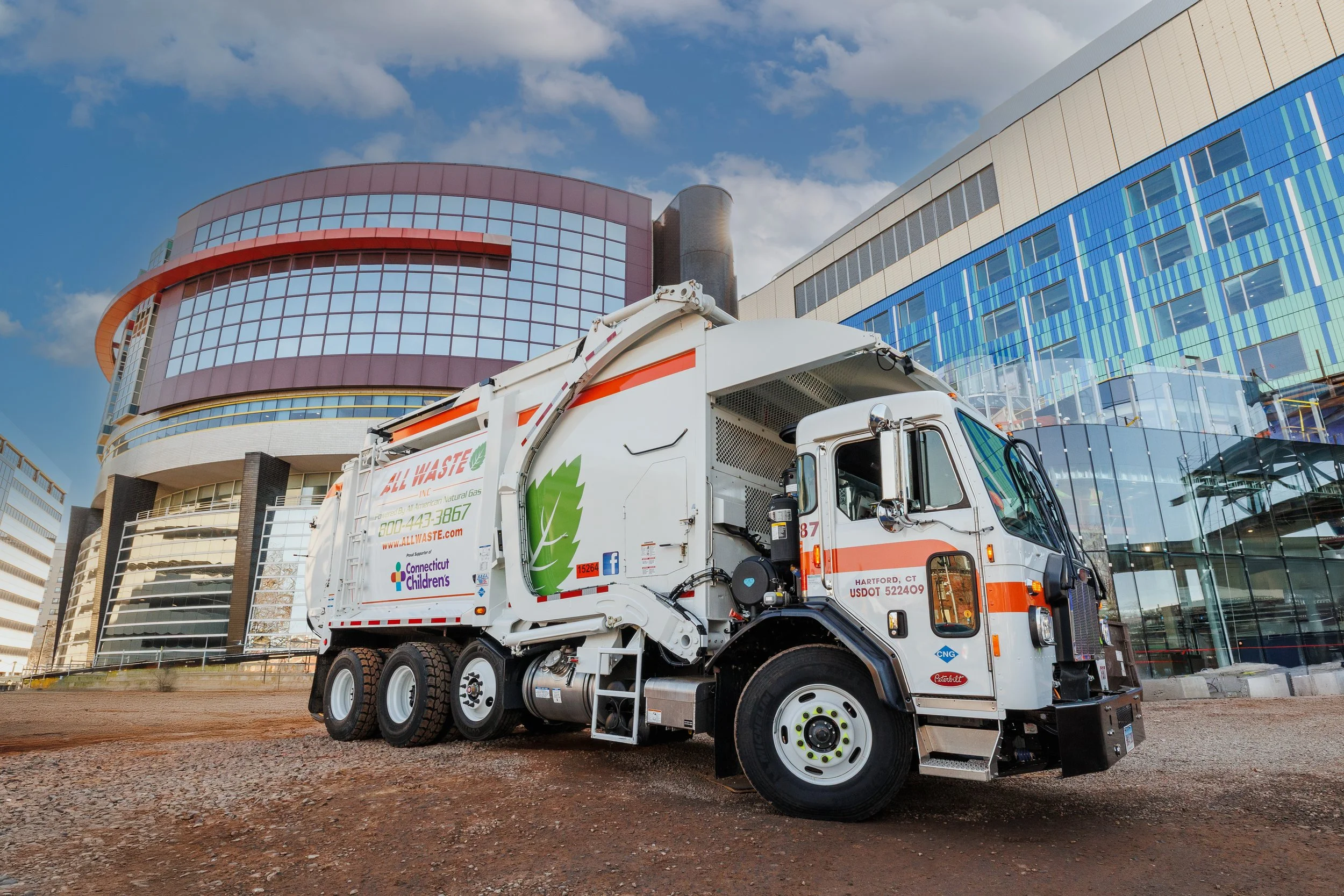 A trash collection truck with Green Leaf logo parked on a dirt surface in front of a modern glass building under a partly cloudy sky.