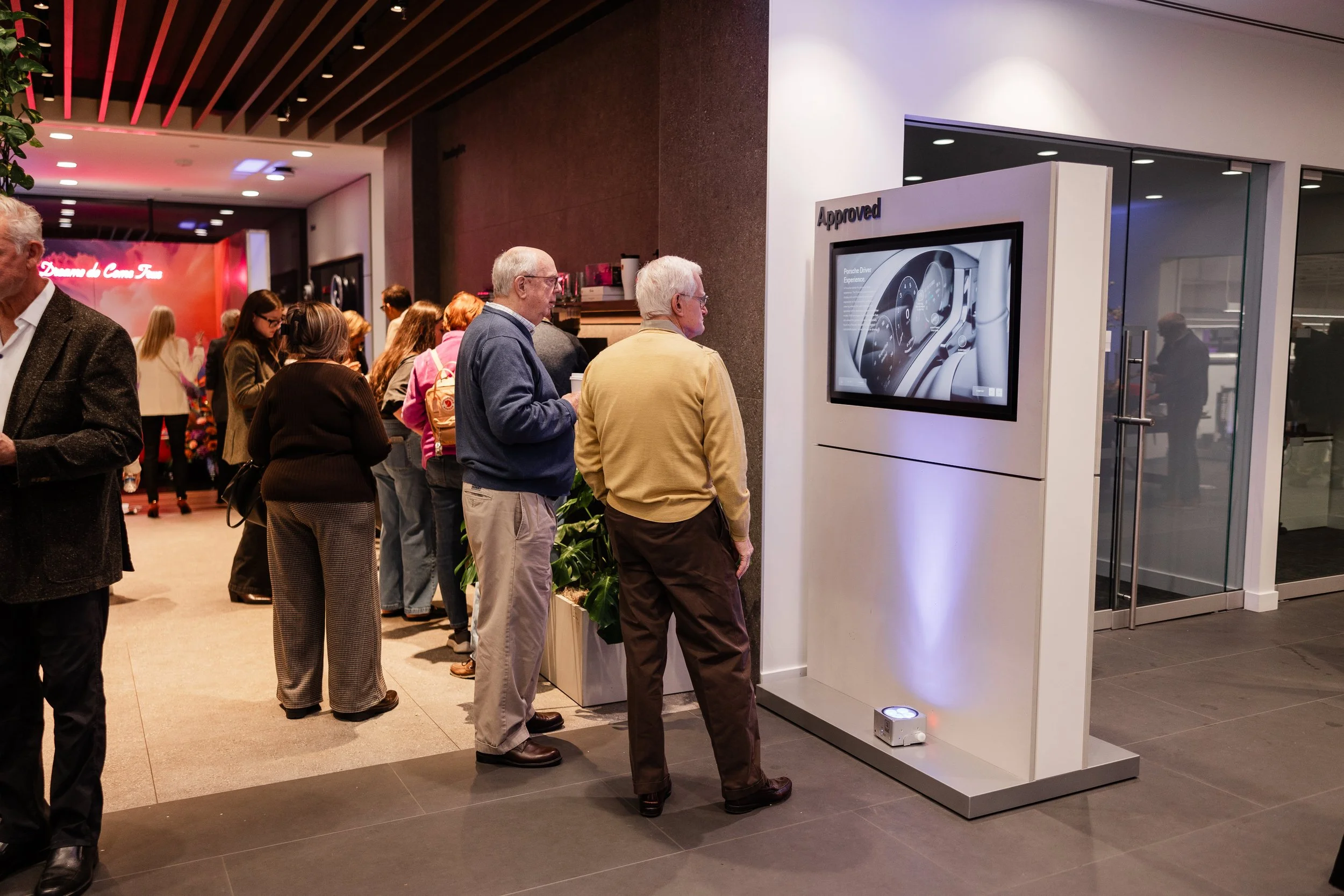 People standing in line at a digital kiosk displaying a car dashboard image, inside a modern building with a glass door entrance.