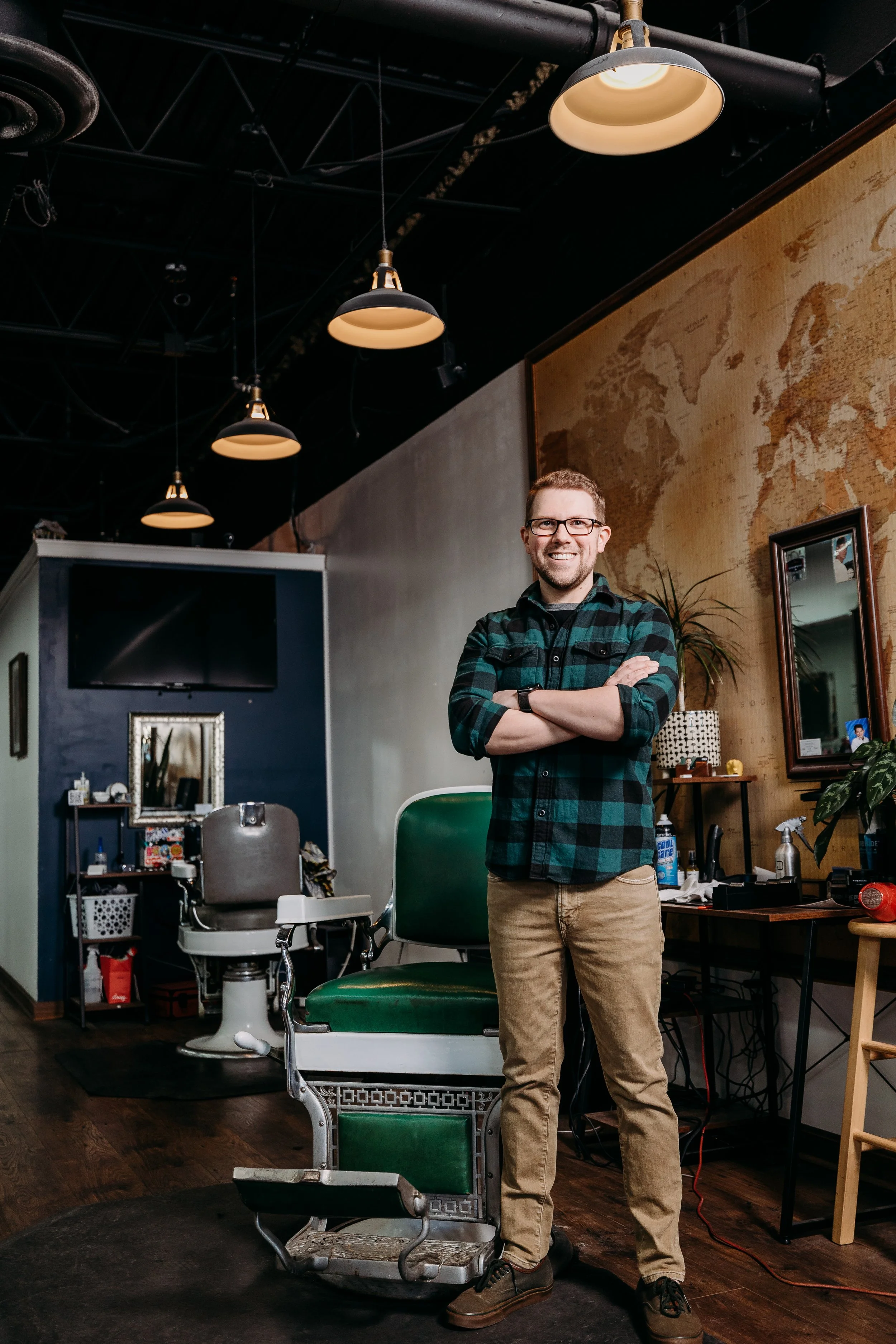 A smiling man with glasses and a beard standing arms crossed in a barbershop or hair salon, with vintage barber chair, mirror, plants, and wooden wall decor around him.