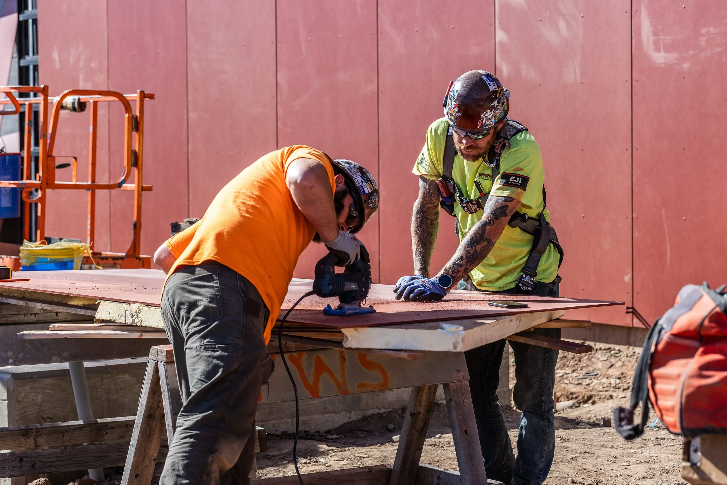 Two construction workers, one in an orange shirt and the other in a yellow safety vest, are working together on a piece of wood at a construction site. They are wearing helmets and gloves, with a pink wall behind them.