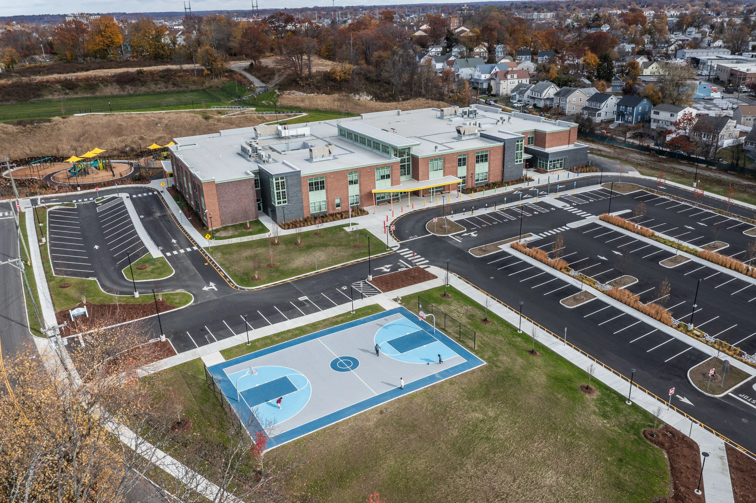 An aerial view of a modern school campus showing a large building, a parking lot, a basketball court, and a playground in a suburban neighborhood with houses and trees in the background.