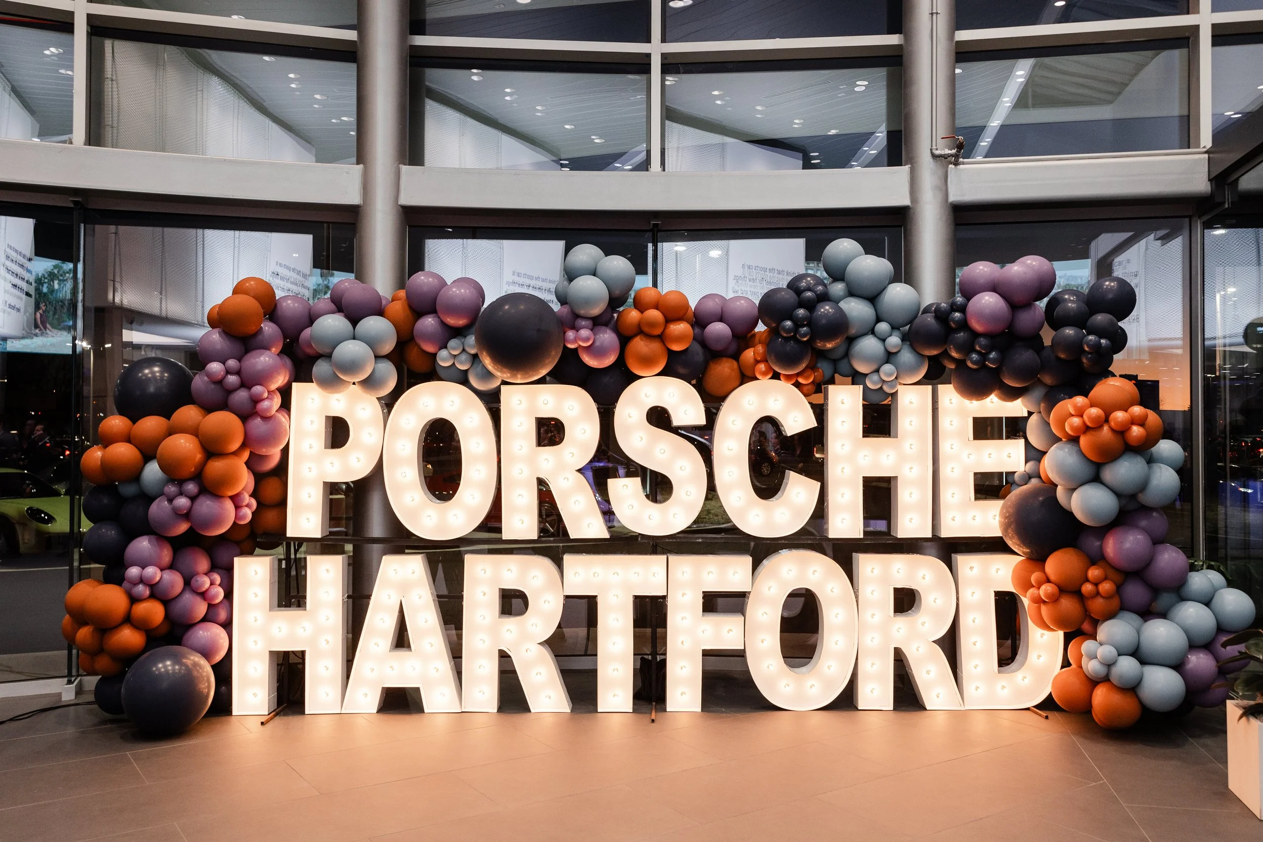Decorative display with large illuminated letters spelling 'Porsche Hartford' surrounded by colorful balloons in purple, orange, black, and light blue at a car dealership