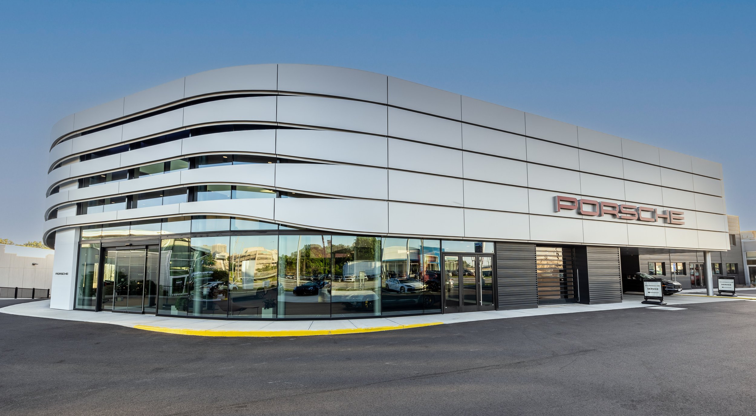 Modern Porsche dealership building with large glass windows and the Porsche logo on the exterior, parked cars inside and outside, and a clear blue sky overhead.
