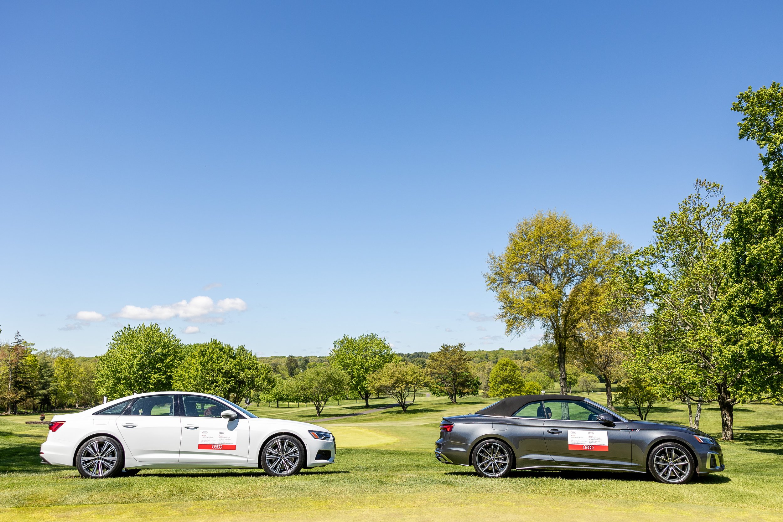Two Audi convertibles, one white and one gray, parked on a lush green golf course under a clear blue sky with trees in the background.