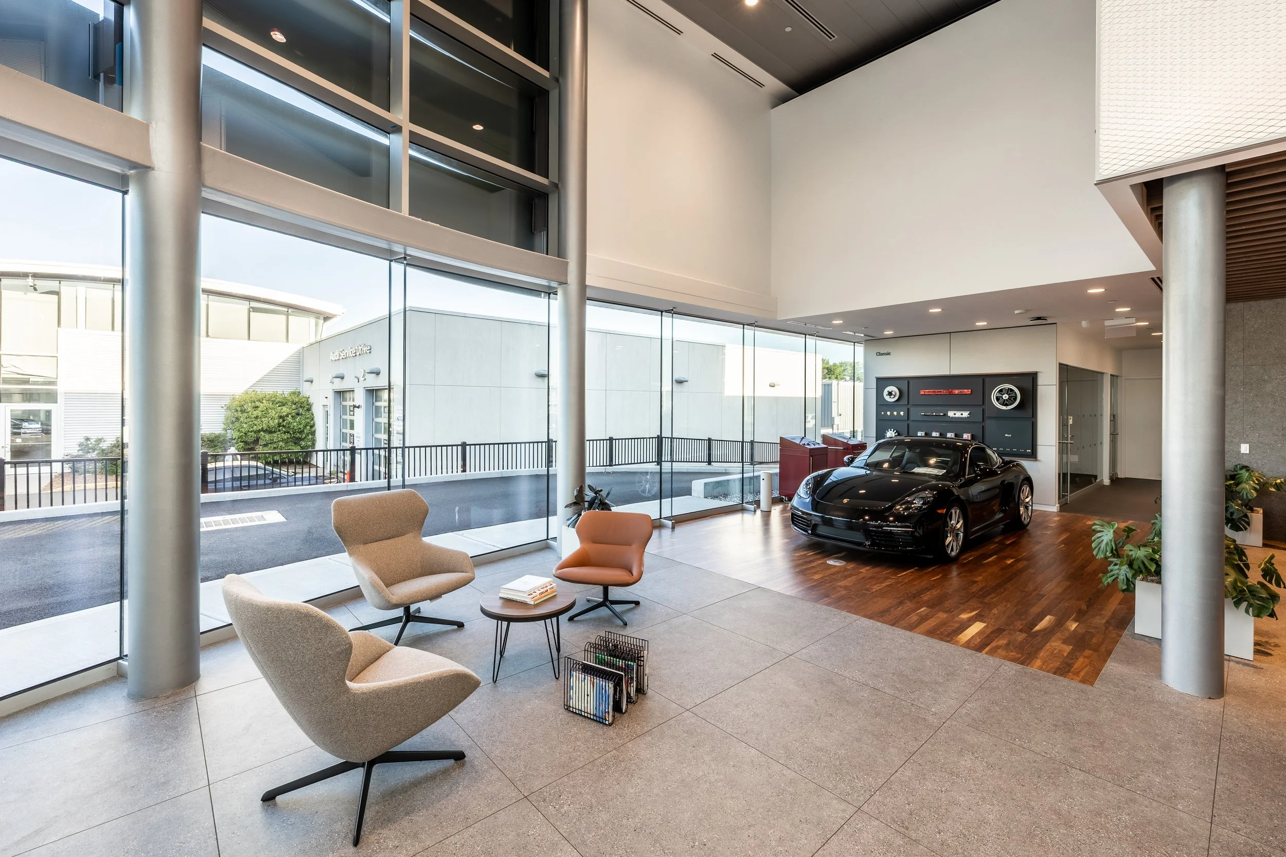 Modern car dealership showroom with large glass windows, a black sports car on a wooden floor, seating area with four chairs, potted plants, and classic car memorabilia on the wall.