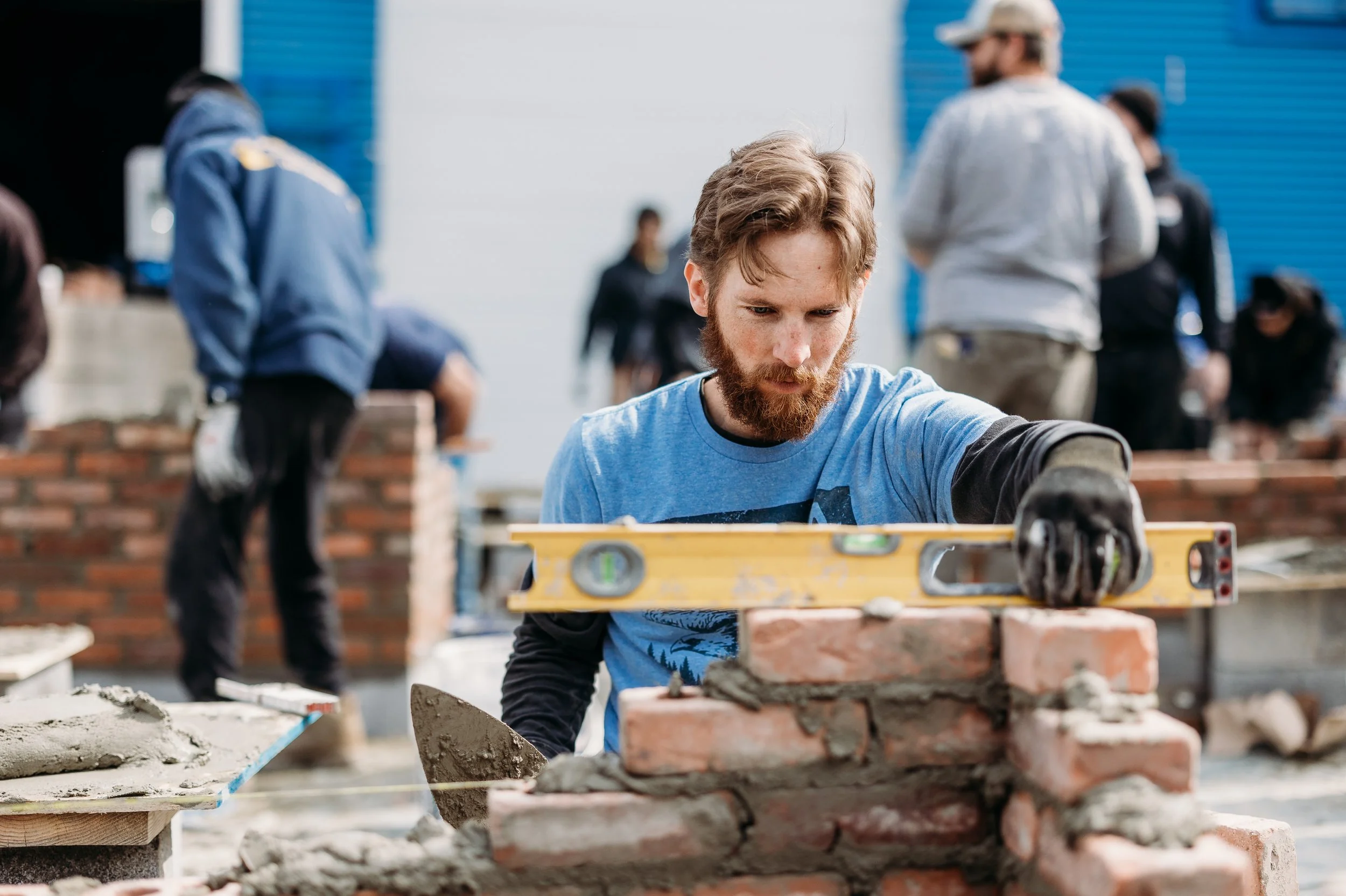 Man with a beard and blue t-shirt building a brick wall, using a spirit level, at a construction site, with several people in the background also working.