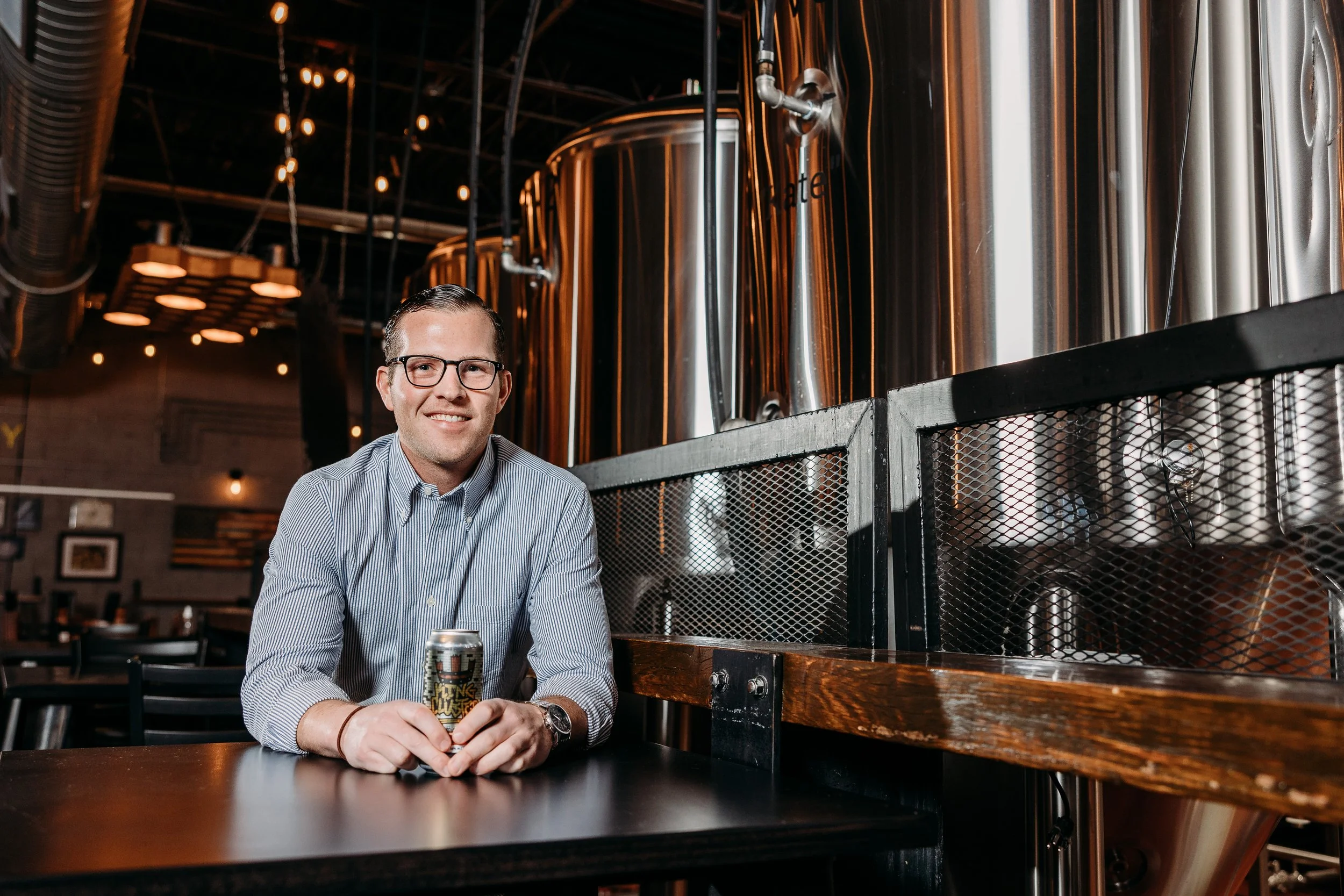 A smiling man with glasses and a gray striped shirt sitting at a table in a brewery, holding a can of beer, with large stainless steel brewing tanks in the background.
