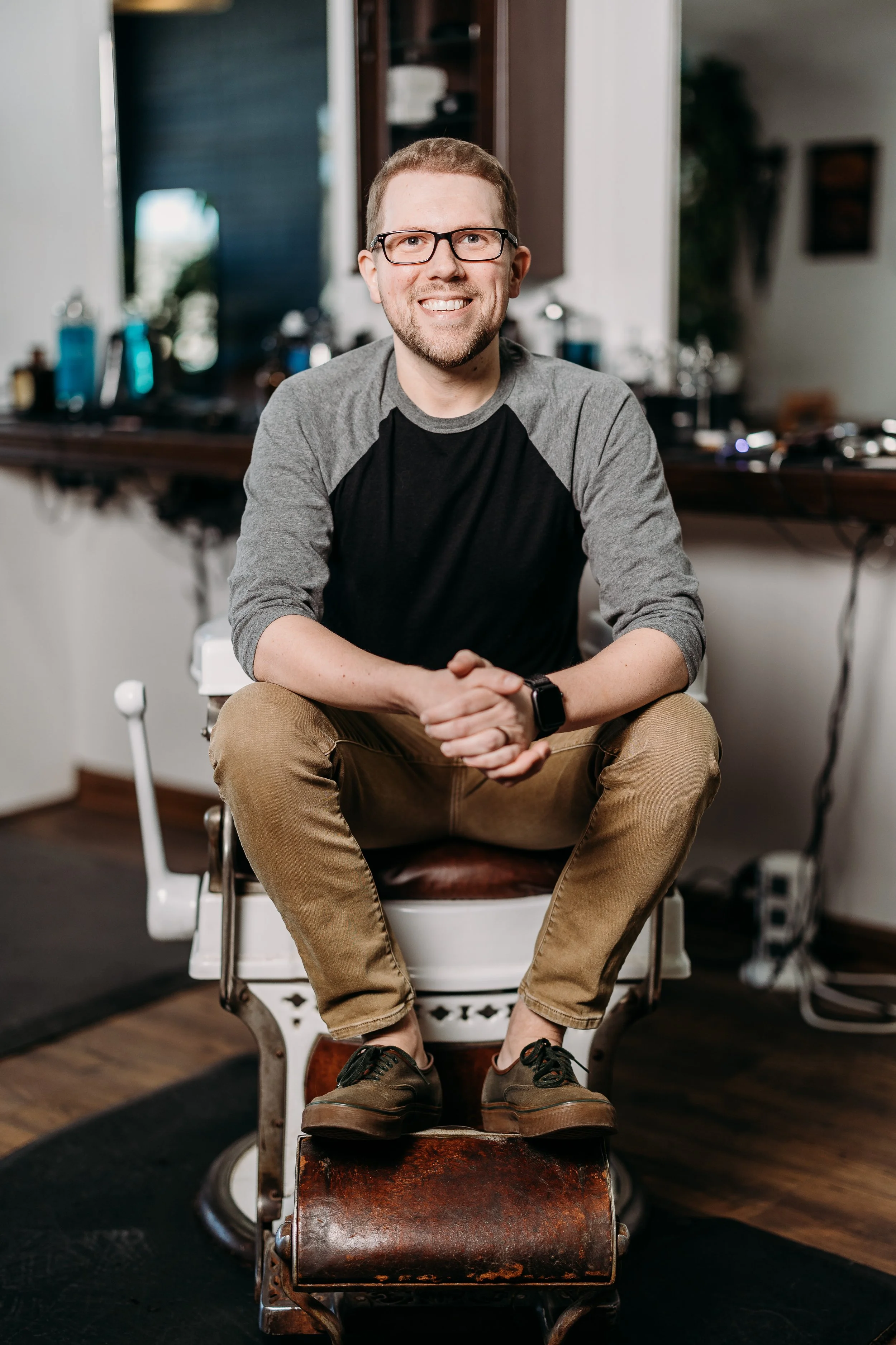 A smiling man with glasses, a beard, and short hair sitting on an old-fashioned barber's chair in a barbershop or salon, with various hair care products and tools visible in the background.
