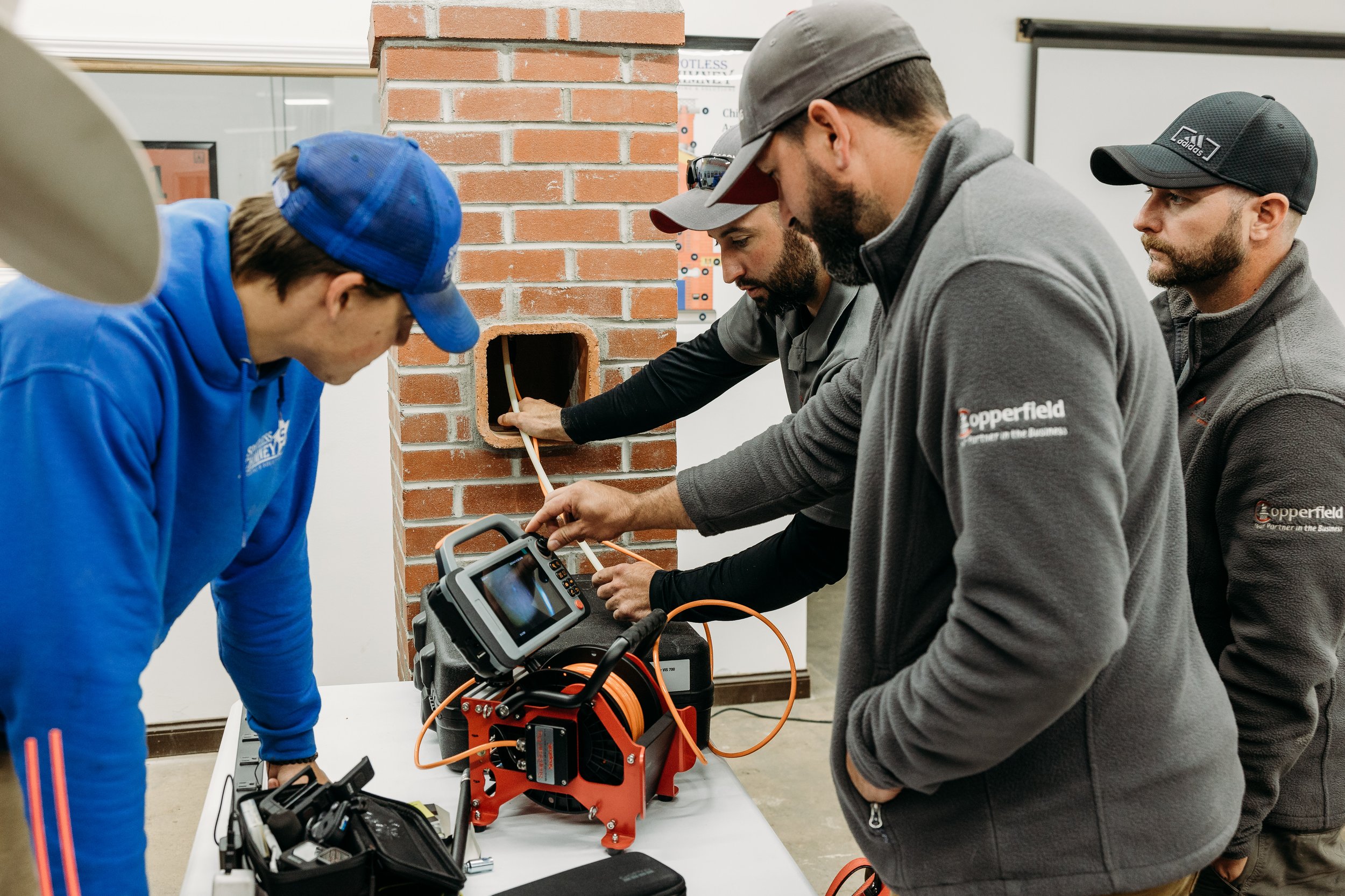 Group of four men working on electrical equipment at a table, one man is reaching into a hole in a brick wall, with tools and a diagnostic device on the table.