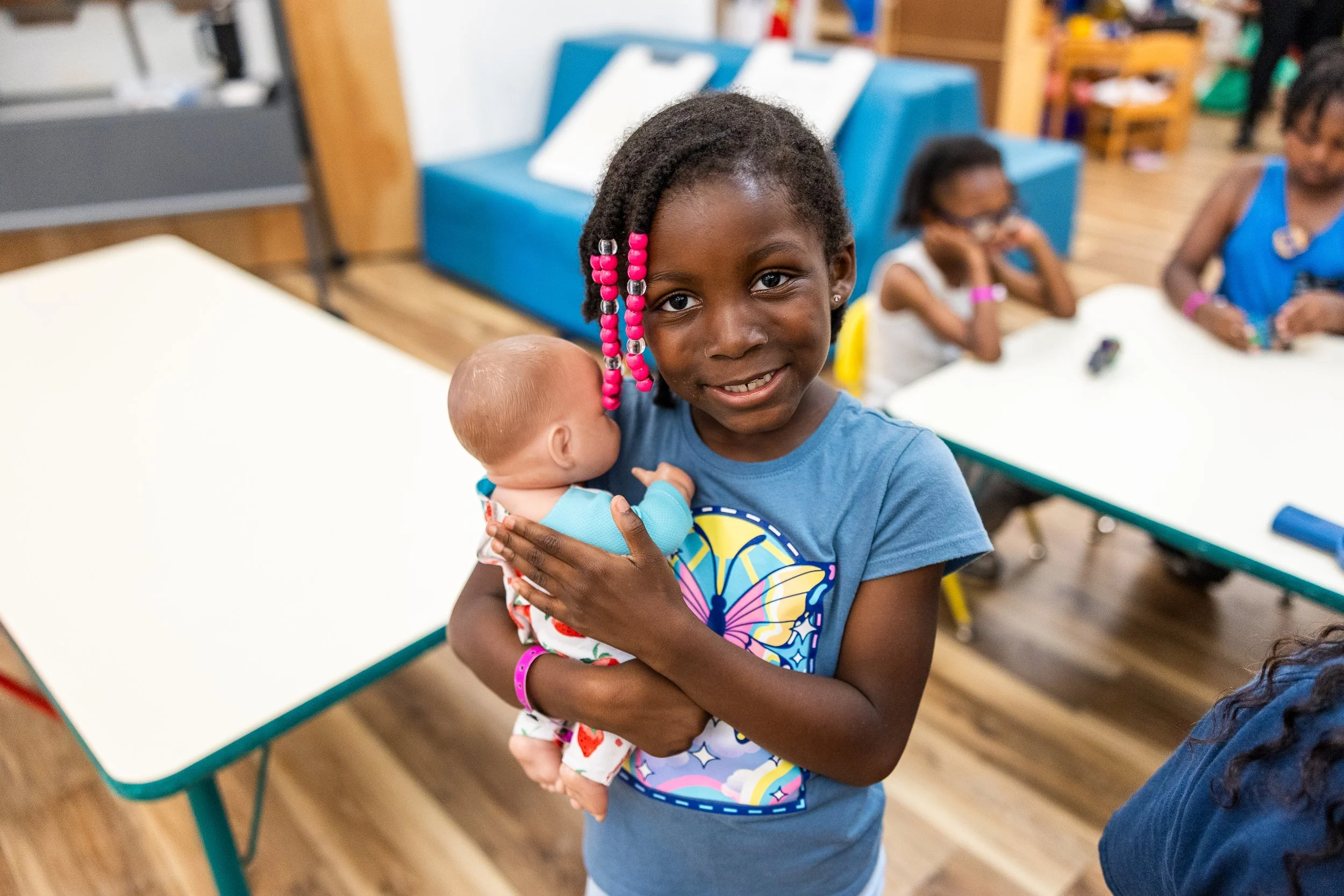 A young Black girl with pink beads in her hair holding a sleeping baby doll, smiling at the camera in a classroom setting with other children in the background.