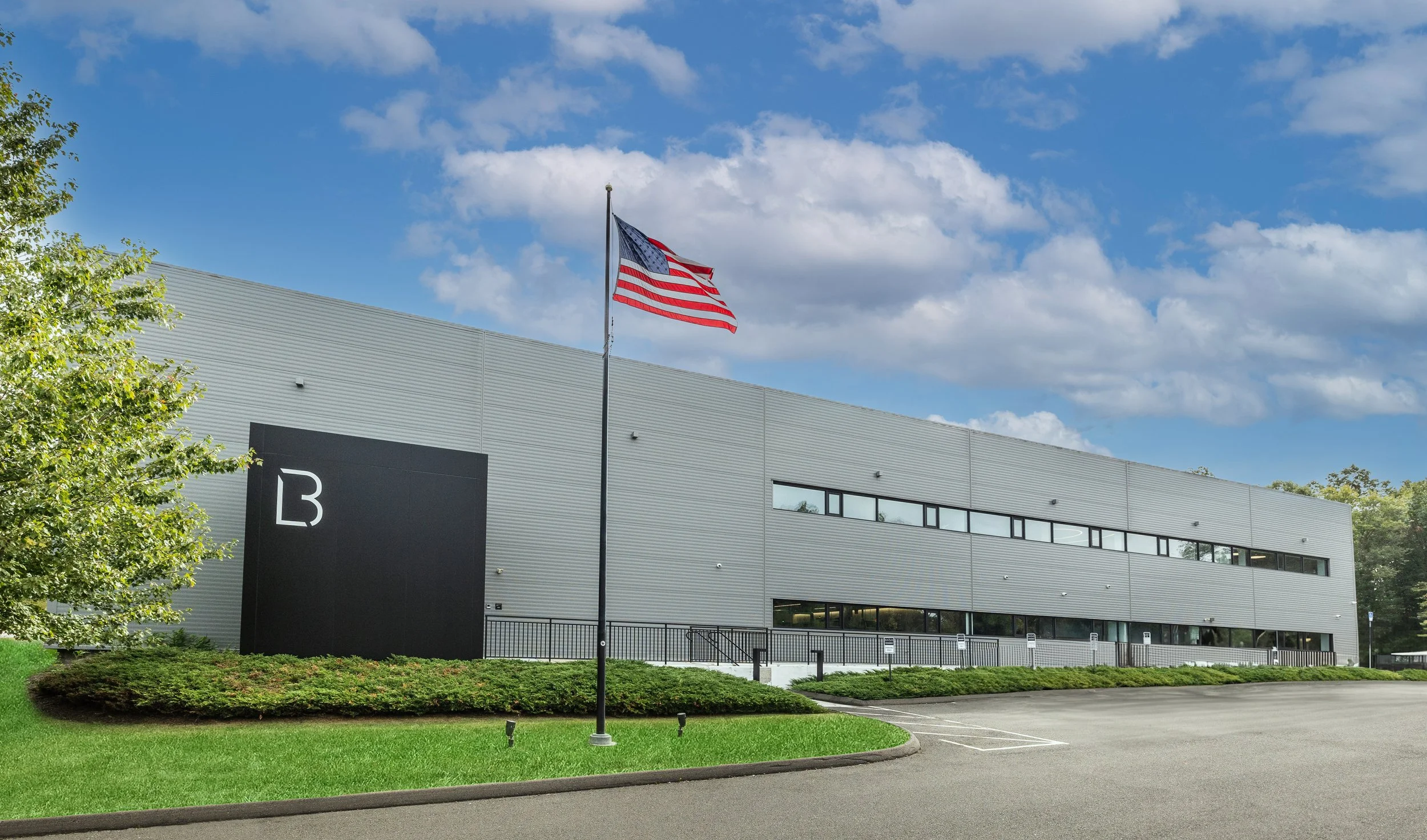 Modern gray industrial building with a black sign displaying a white 'B', a flagpole with an American flag, a parking lot, and a partly cloudy sky.