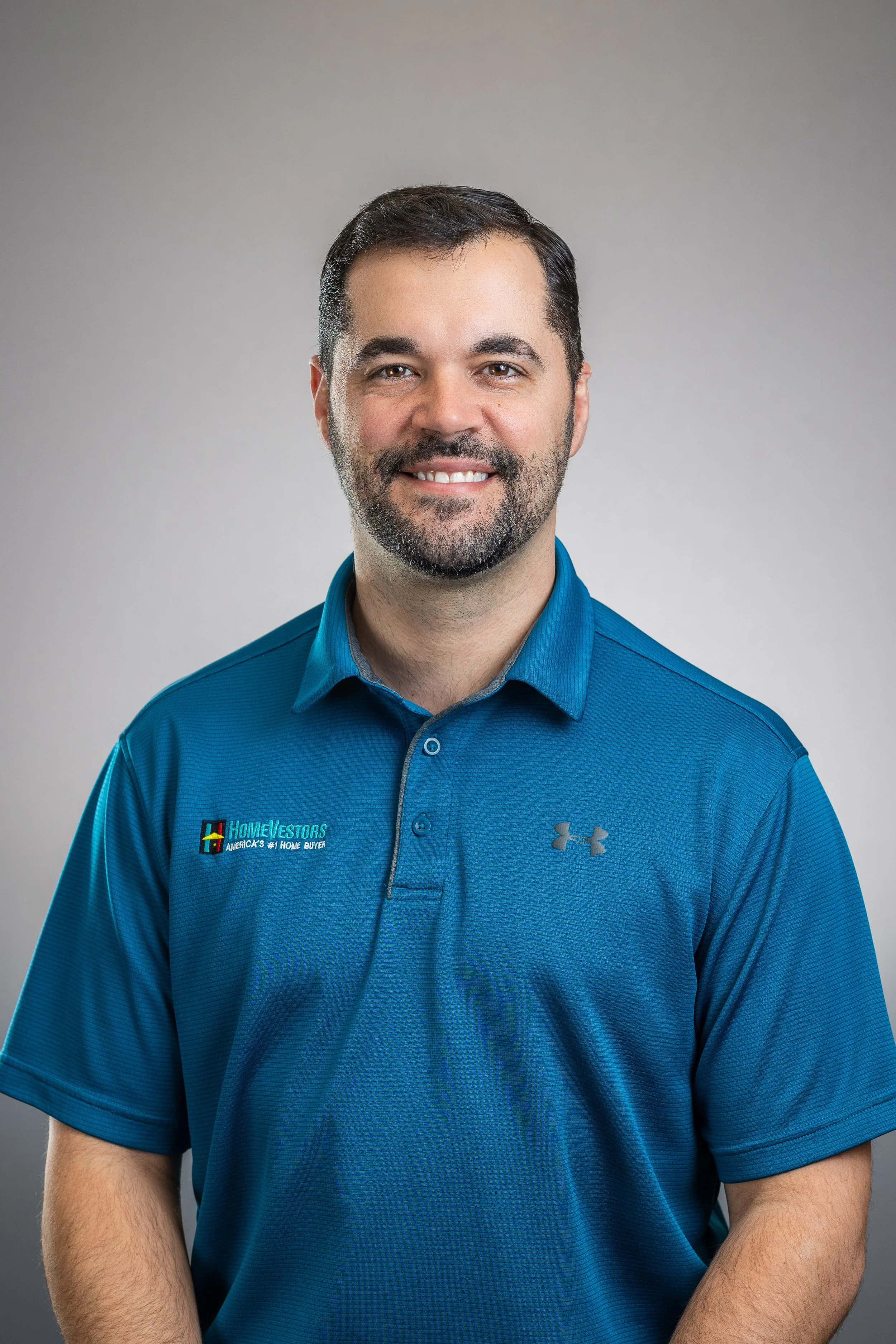 A man with short dark hair and a beard, smiling, wearing a blue polo shirt with a logo and text, standing against a gray background.
