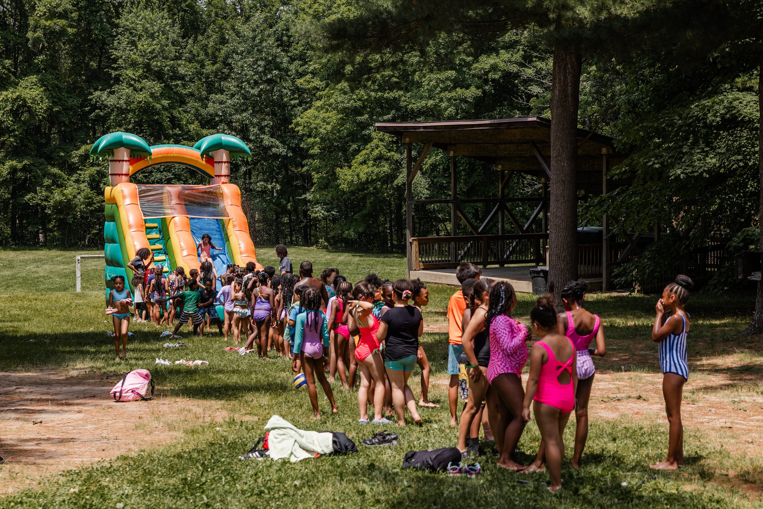 Group of children in swimsuits waiting in line for a water slide at an outdoor park, with lush green trees in the background.