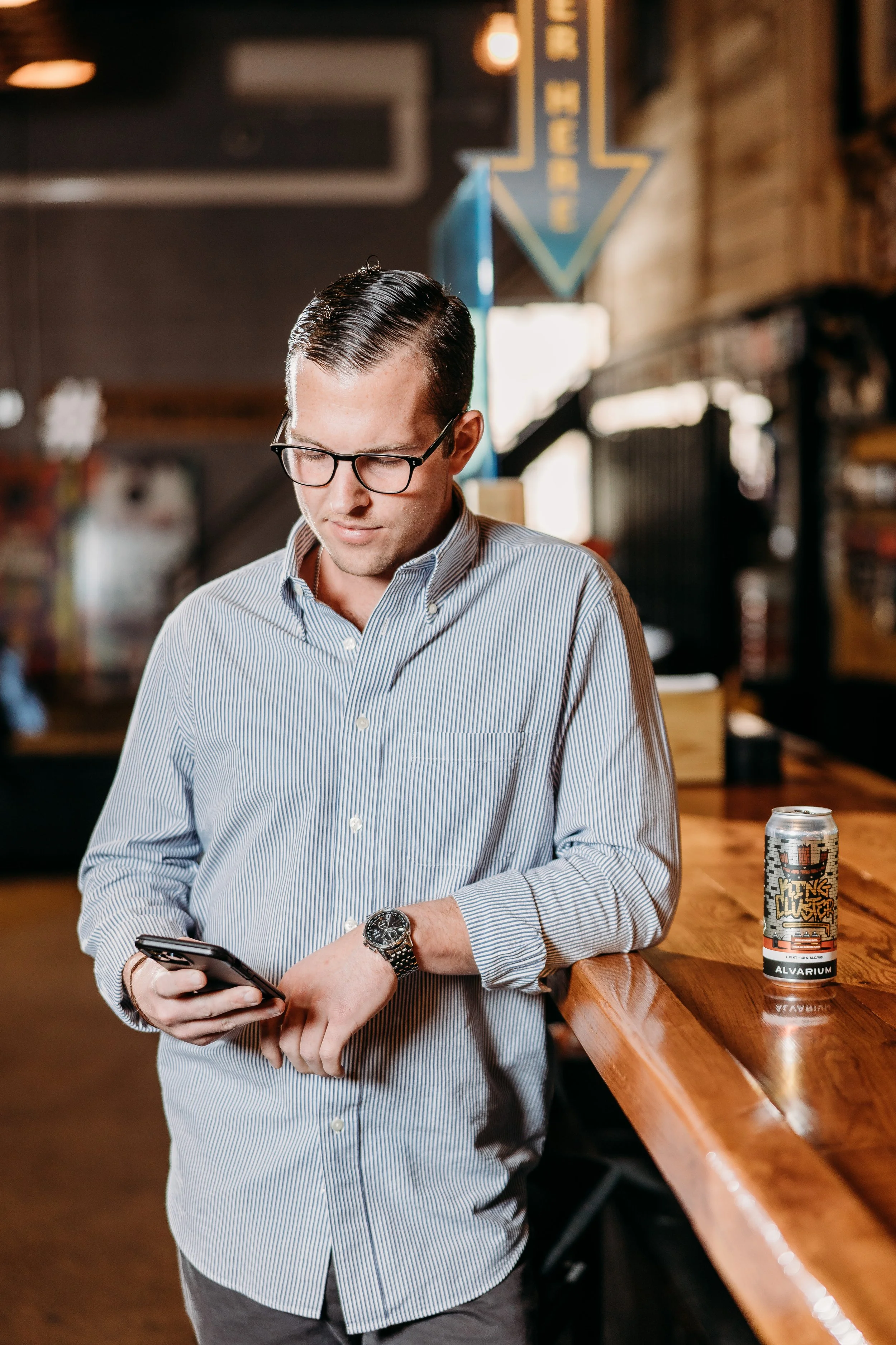 A man in a striped button-up shirt and glasses standing at a wooden bar, looking at his phone. A can of Alvarium beer is on the bar.