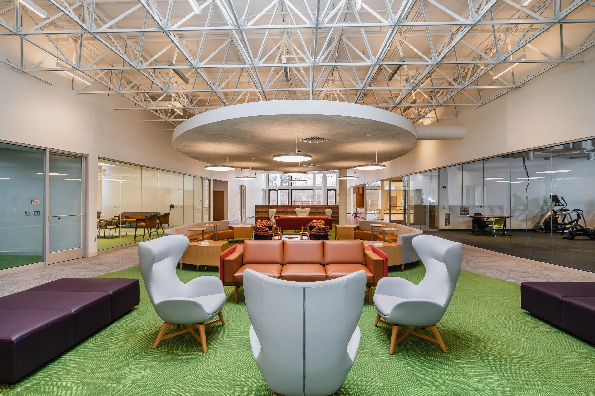 Modern indoor lounge area with colorful seating, green carpet, glass walls, and high ceiling with exposed beams and circular light fixtures.
