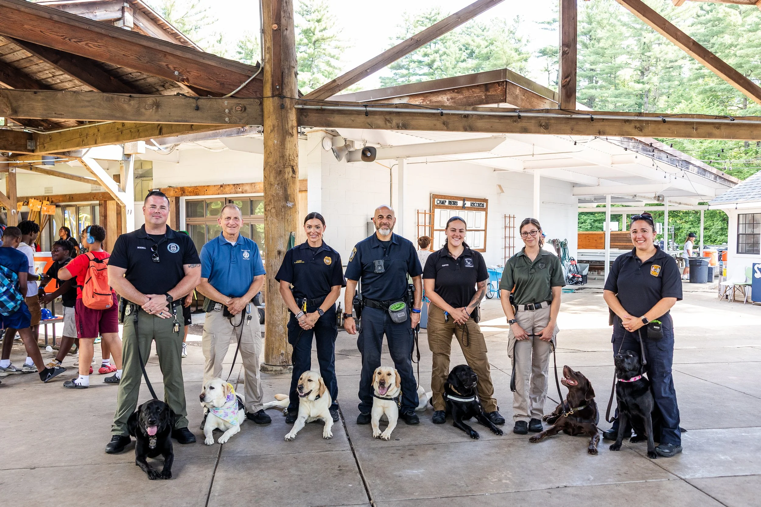 Group of seven uniformed police officers with service dogs standing in a pavilion at a camp.