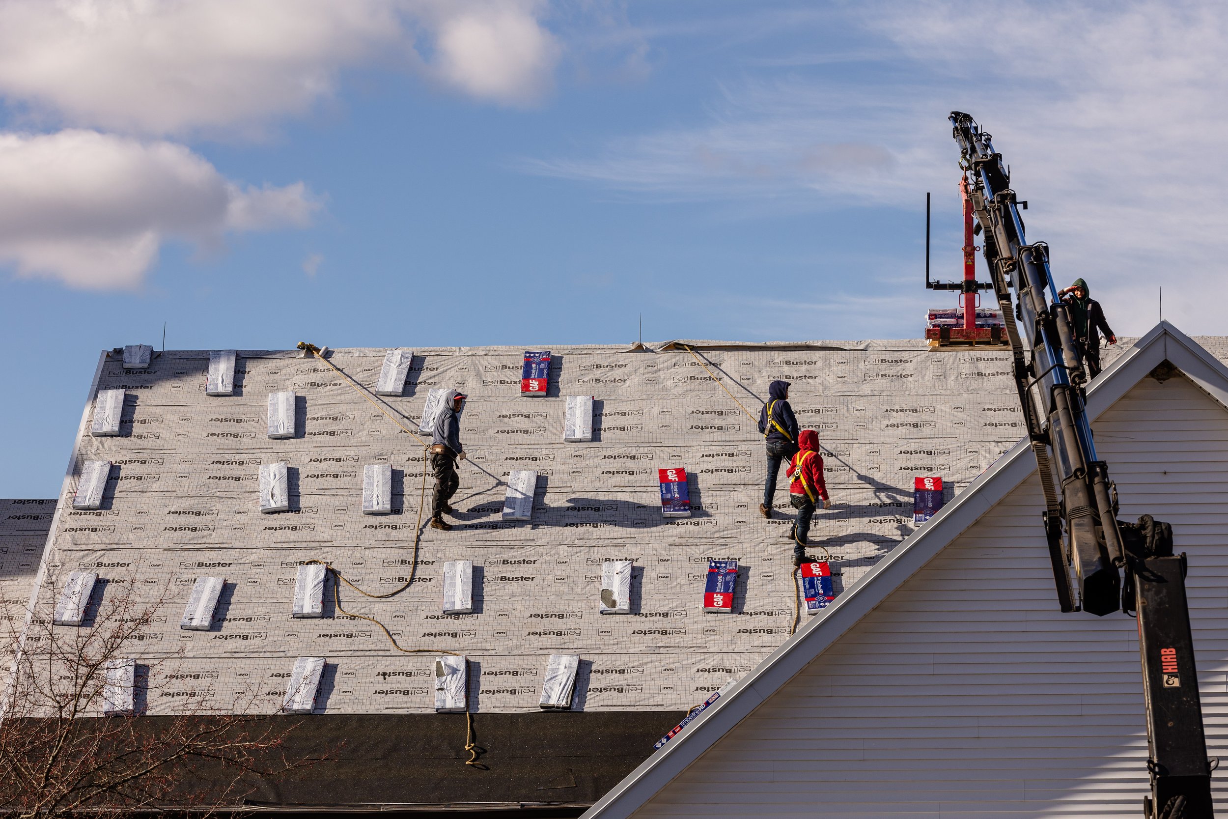 Construction workers installing shingles on a house roof, with a crane on the side.