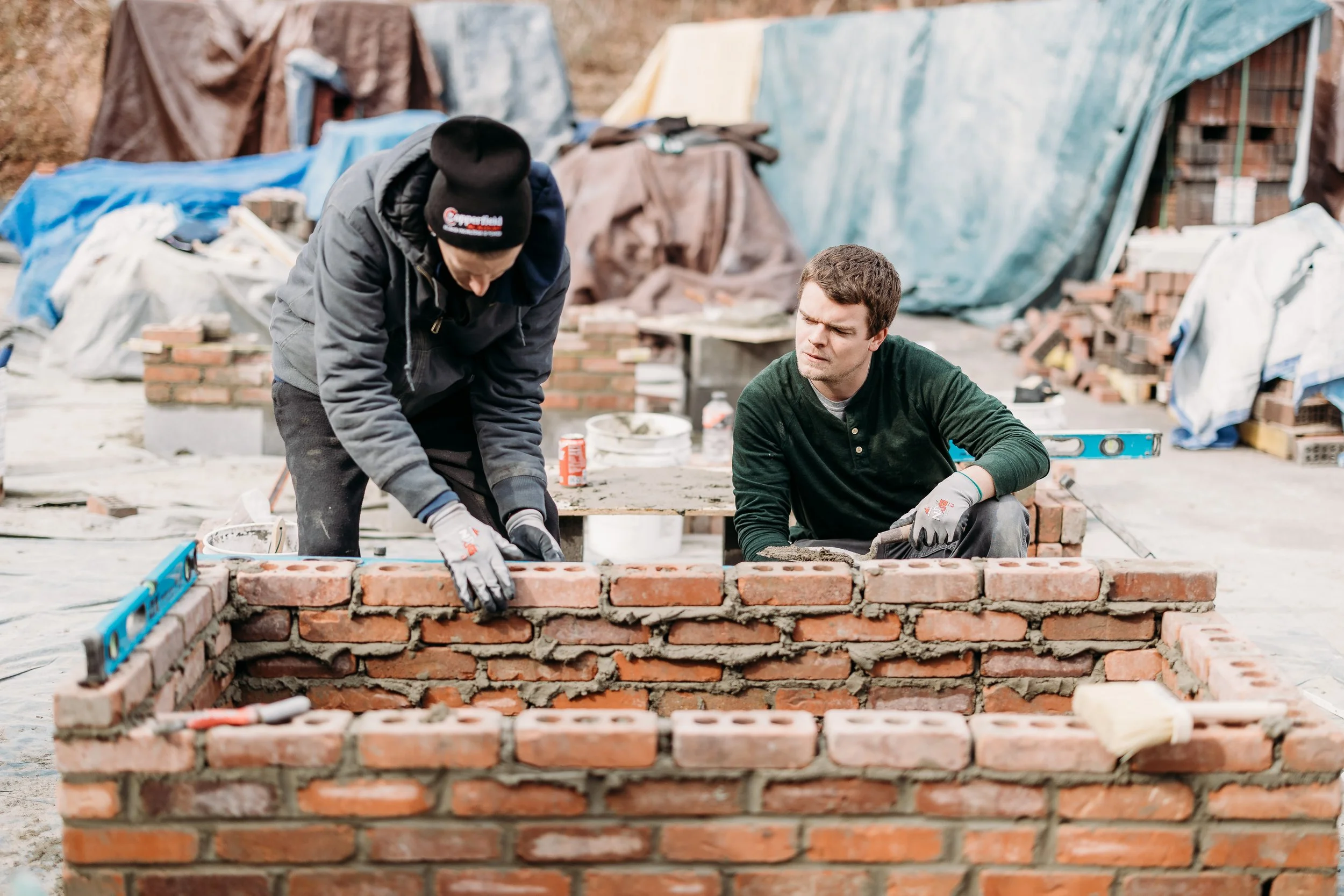 Two men building a brick wall outdoors on a construction site, with construction materials and tents in the background.