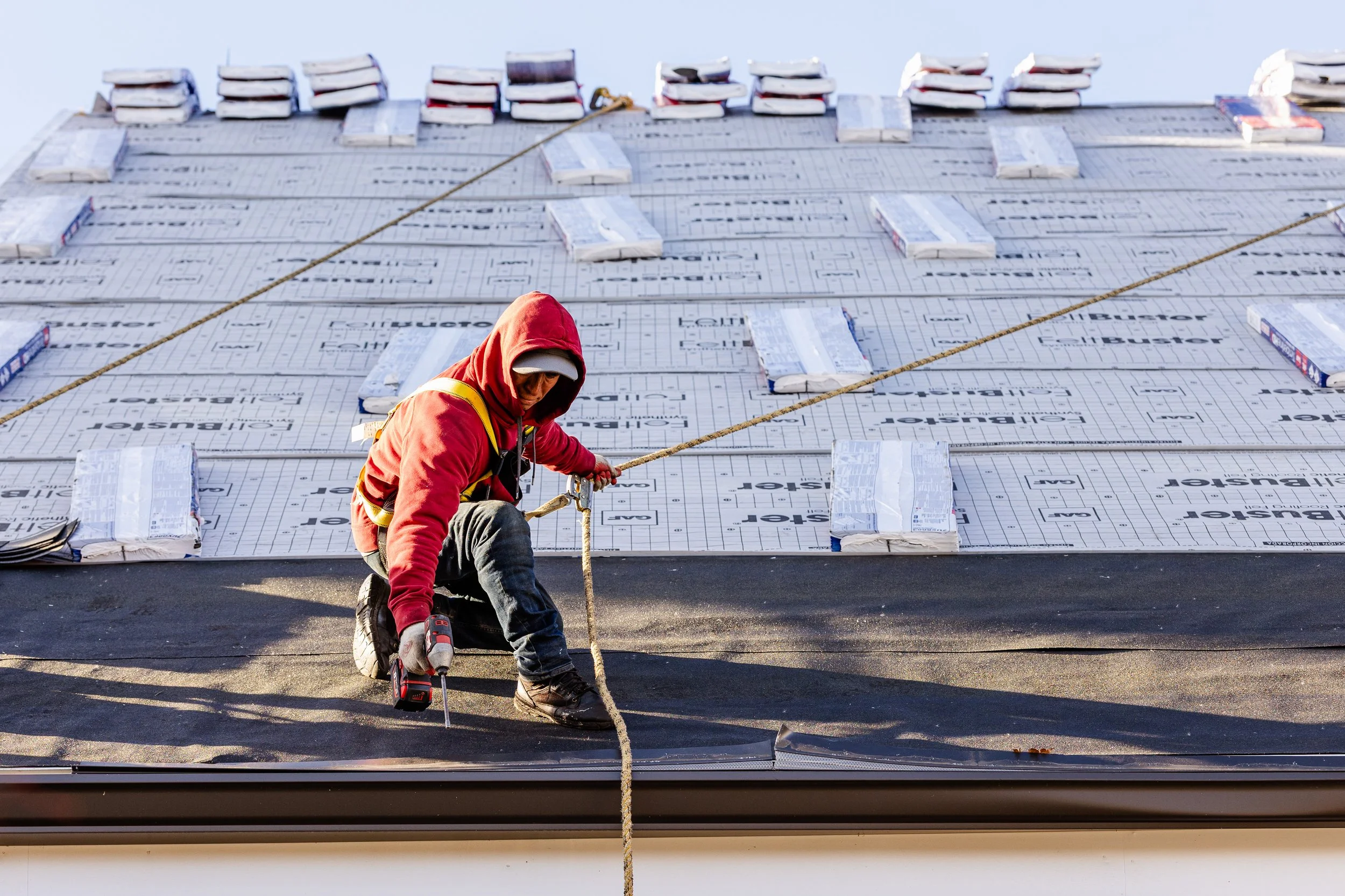 A construction worker is installing a rubber membrane on a roof. The worker is wearing a red hoodie, safety harness, and gloves, holding a drill. Several packs of roofing material are laid out on the roof surface, and a rope is attached for safety.