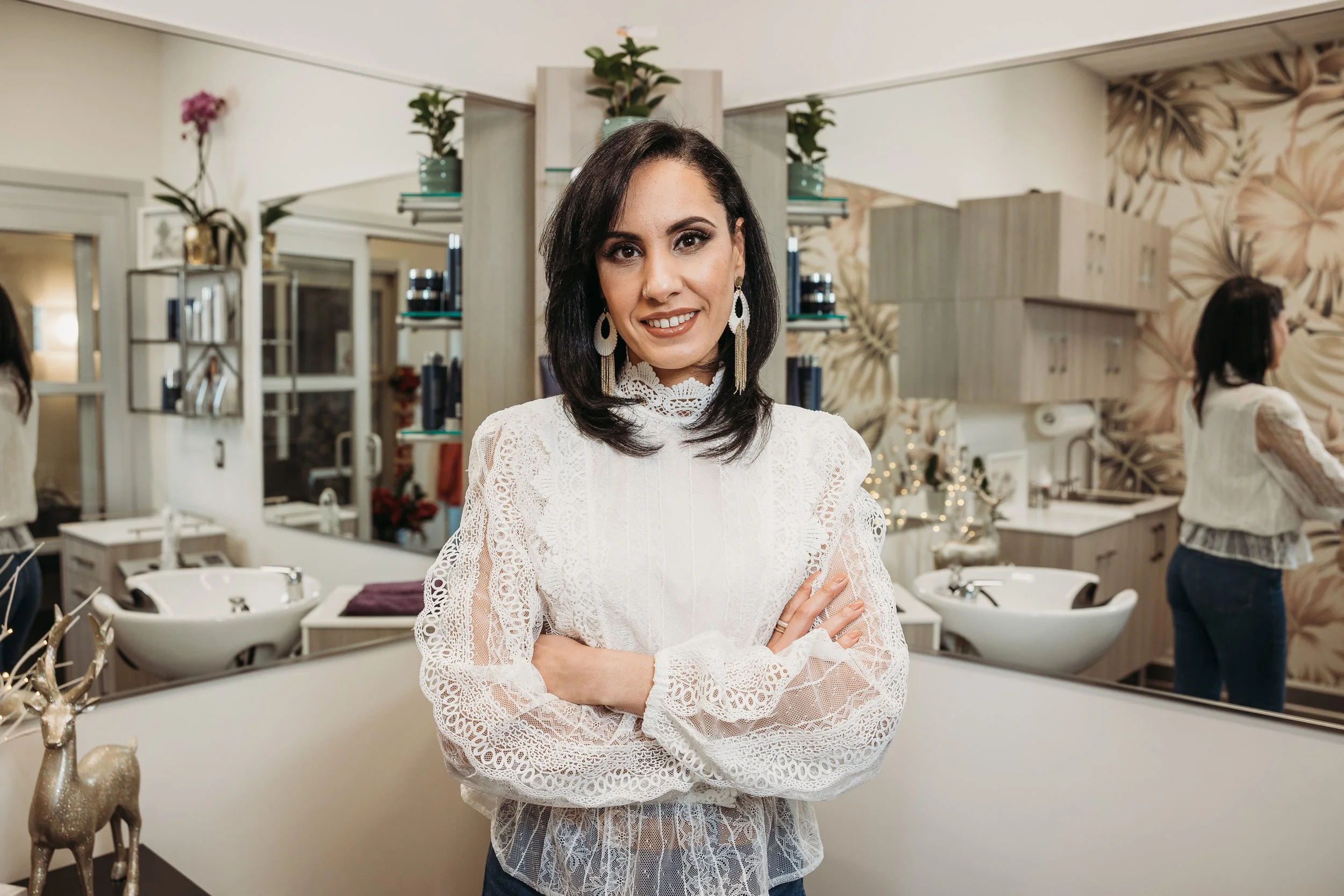 A woman with dark hair wearing a white lace top and large earrings, standing with arms crossed in a decorated salon or beauty studio.