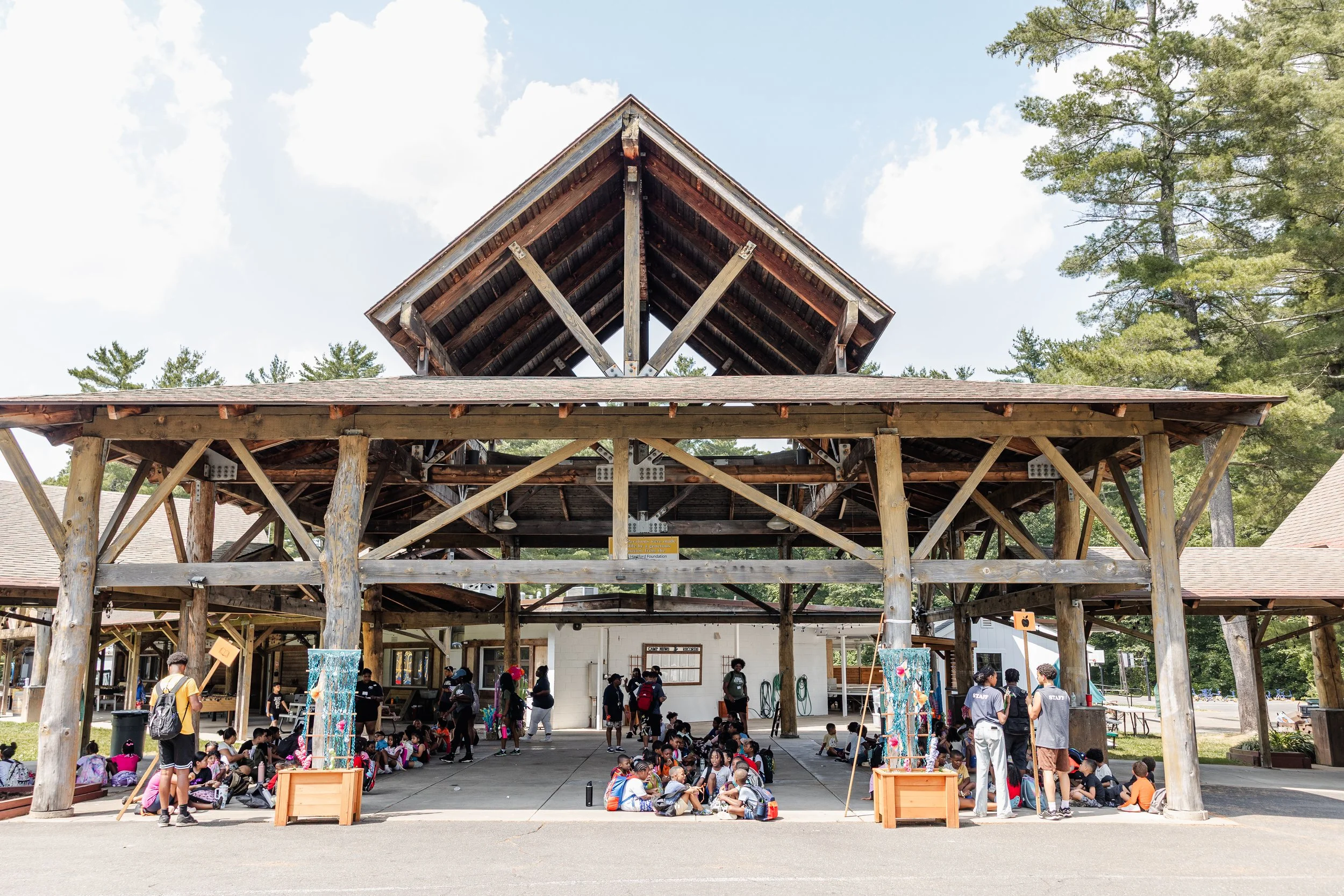 A wooden pavilion with a high sloped roof on a school campus. Children and adults are sitting on the ground beneath the shelter, some students with backpacks, and teachers or staff standing nearby.