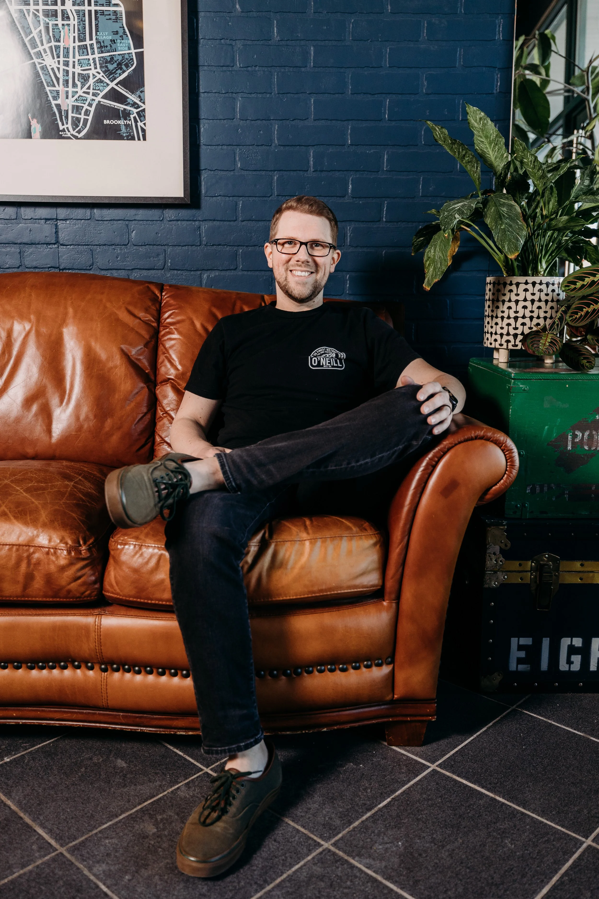 A man sitting on a brown leather couch with his legs crossed, smiling, wearing glasses, a black t-shirt, and dark jeans, with a green plant and a framed map of Brooklyn on a dark blue brick wall behind him.