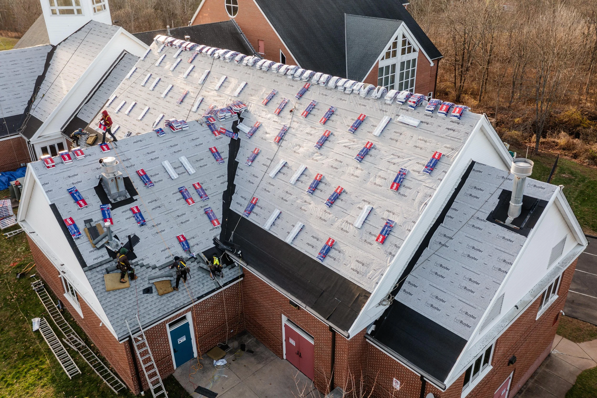 Workers installing roofing on a multi-section brick building, with shingles in progress and roofing materials on the roof.