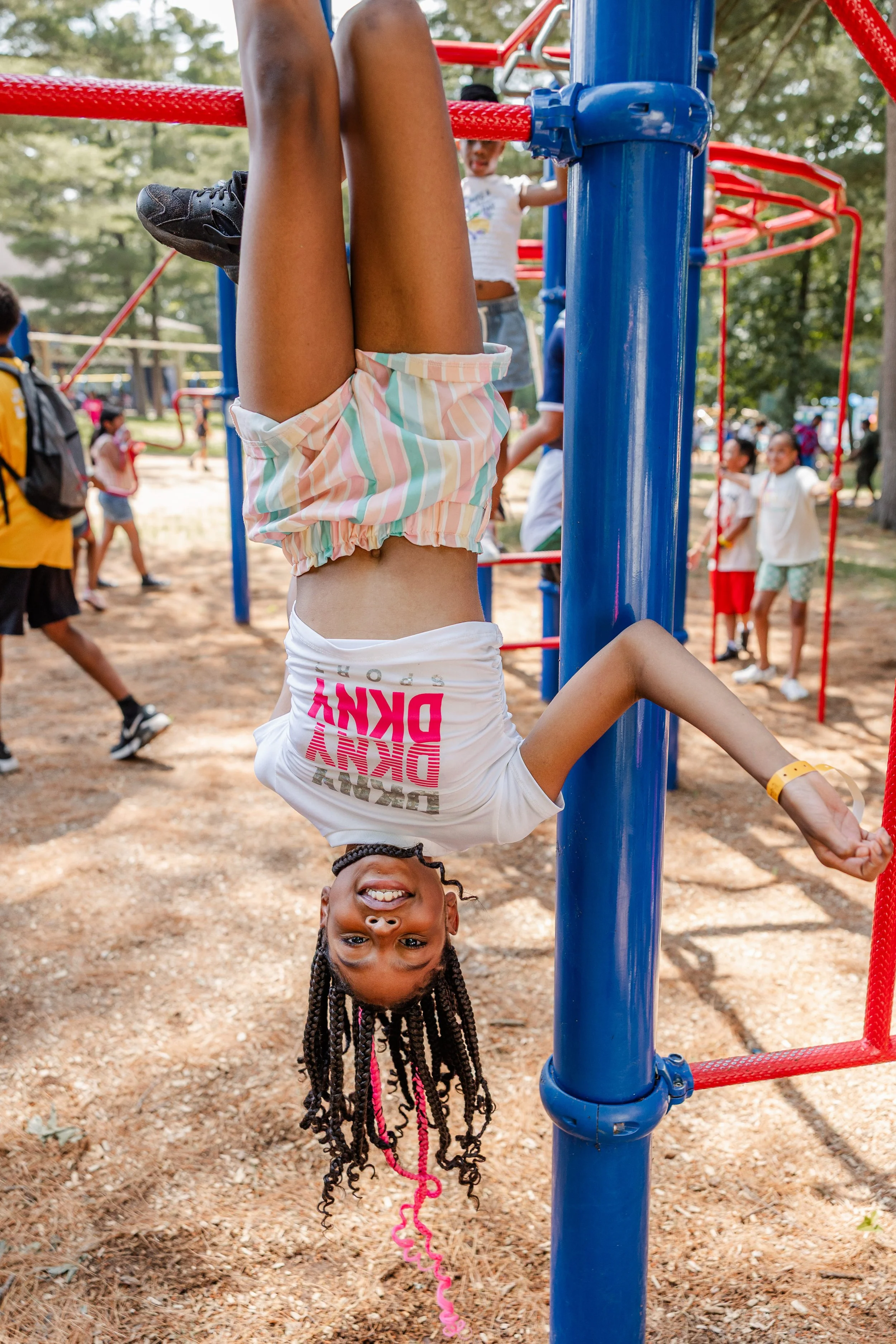 A girl hanging upside down on playground equipment, smiling at the camera, with children playing in the background at a park.
