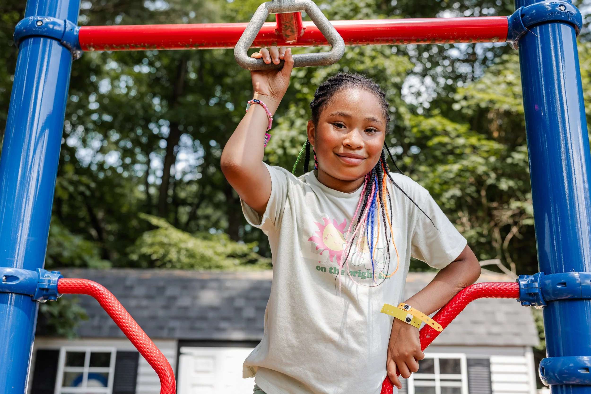 A young girl with colorful braids posing on a playground climbing structure, smiling at the camera, surrounded by trees and a house in the background.