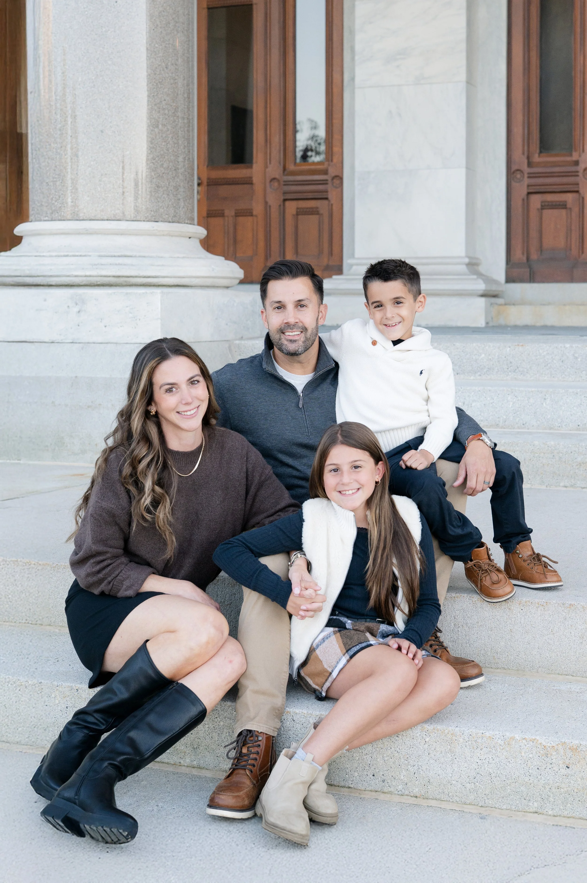 A family of four sitting on the steps in front of a building with large wooden doors. The mother has long wavy brown hair and is wearing a brown sweater, black skirt, and tall black boots. The father has short dark hair, a beard, and is wearing a gray zip-up sweater and beige pants. The son has short black hair and is wearing a white hoodie and dark pants. The daughter has long brown hair and is wearing a plaid skirt, black top, white vest, and beige boots. All are smiling at the camera.